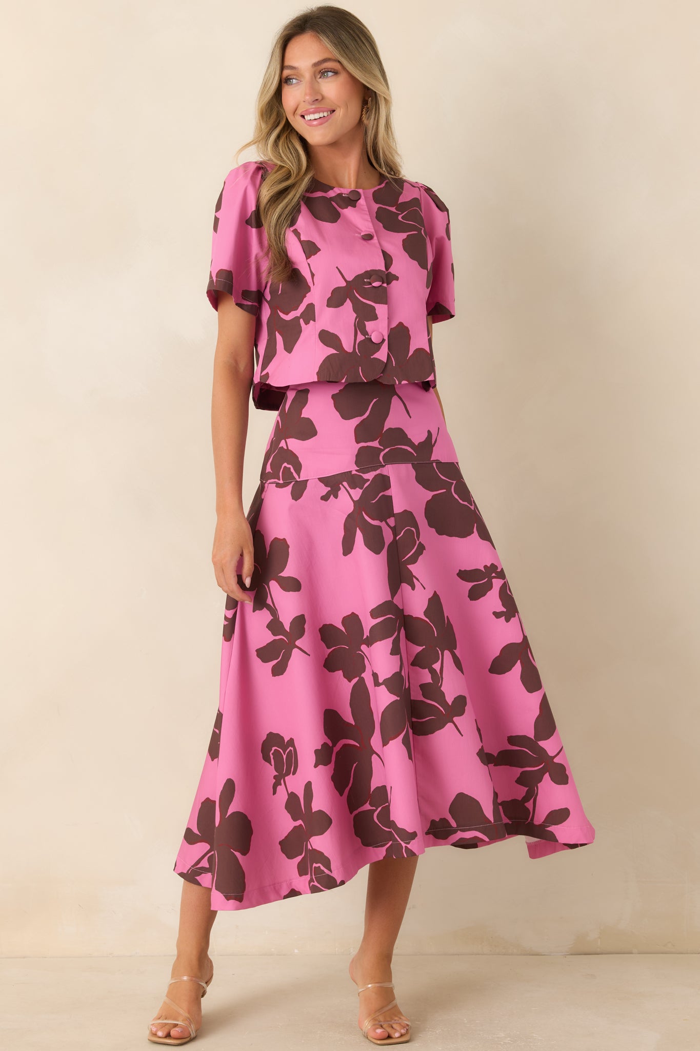 Short-sleeve top featuring a bold chocolate brown floral pattern over bright pink fabric, with a rounded neckline and polished button-front design.