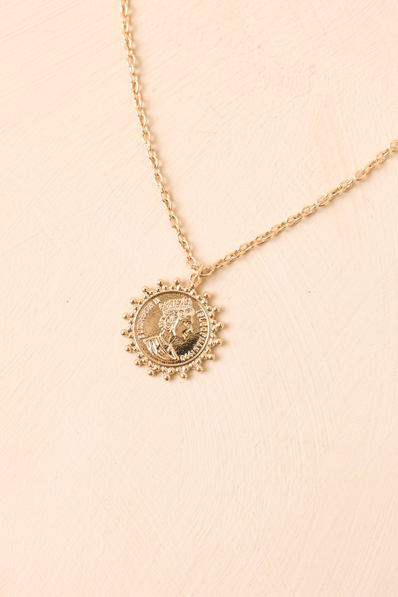 Classic gold necklace against a beige background featuring a framed coin pendant, delicate beaded border, and refined chain design.