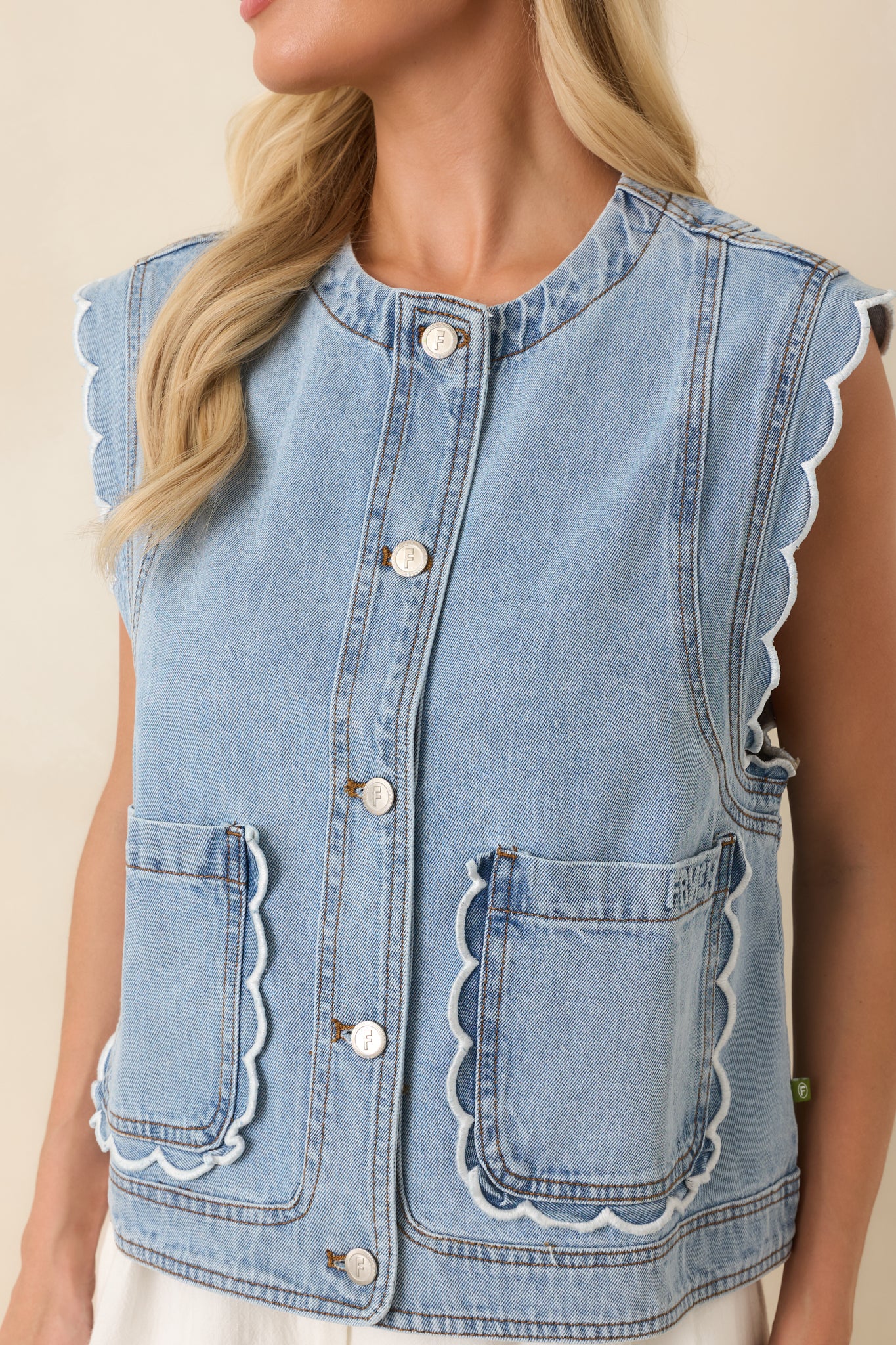 Close-up of the light wash denim vest showcasing the button-front closure, scalloped ruffle pocket trim, textured cotton fabric, and neat stitching.