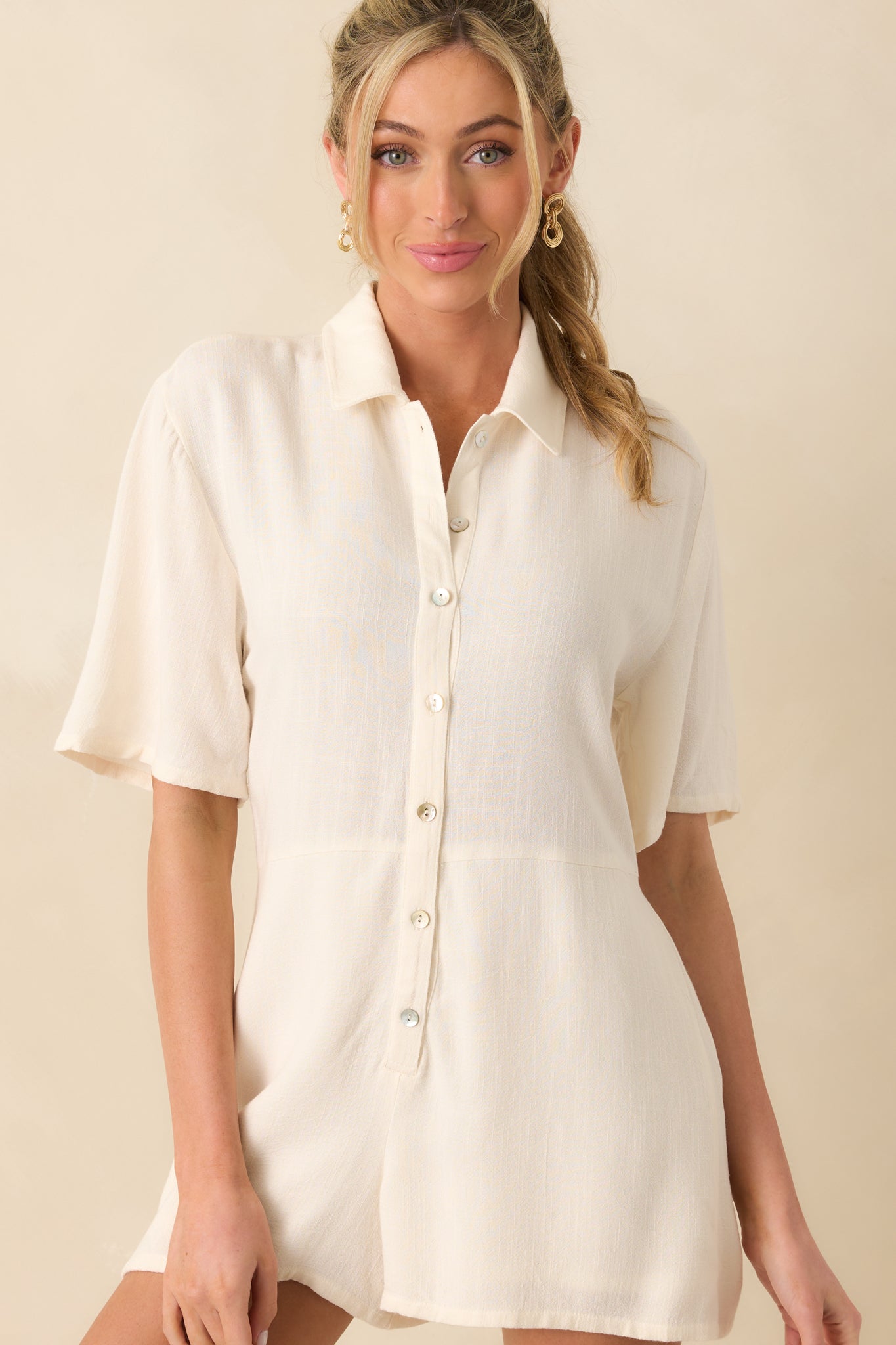 Up-close photo of model wearing ivory romper that features a lightweight rayon-linen blend fabric, collared neckline, functional button front, and short sleeves.