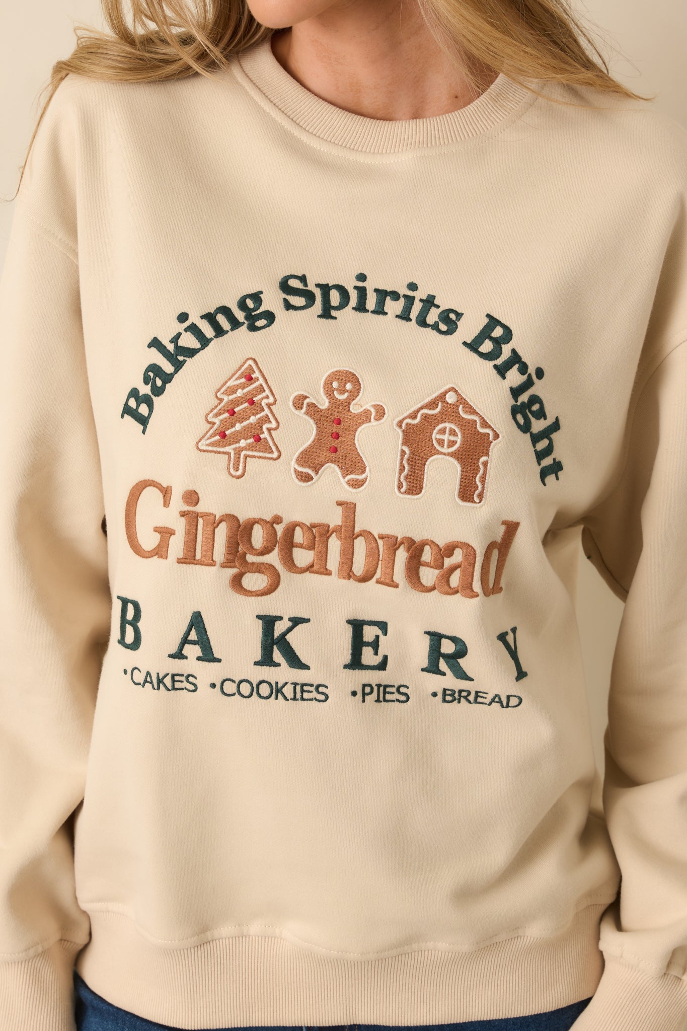 Close-up of the light beige fleece sweatshirt showing the raised gingerbread bakery embroidery and knit texture.