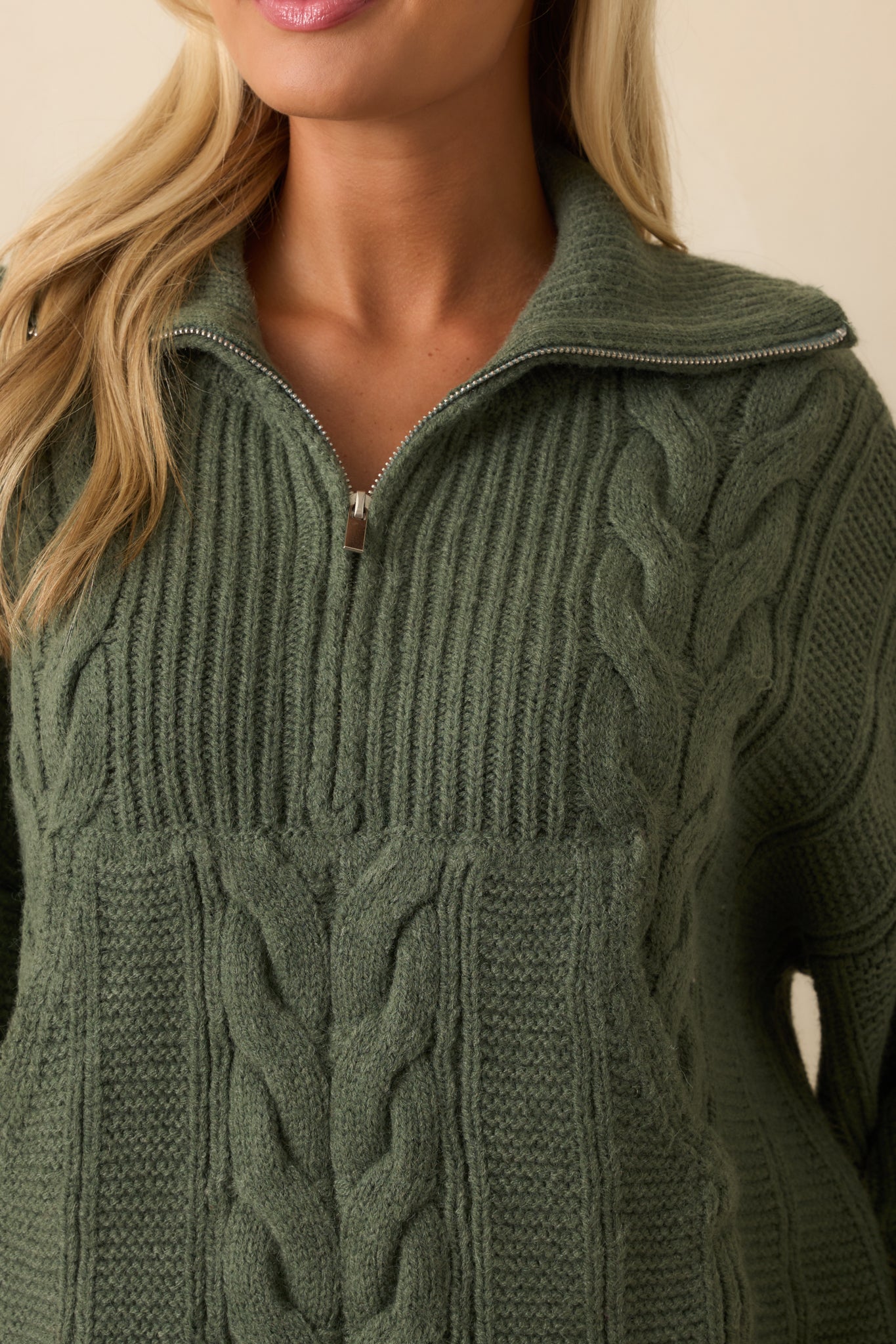 A close-up of the sage green cable knit sweater, showing the knit texture, quarter zip hardware, and ribbed edge.