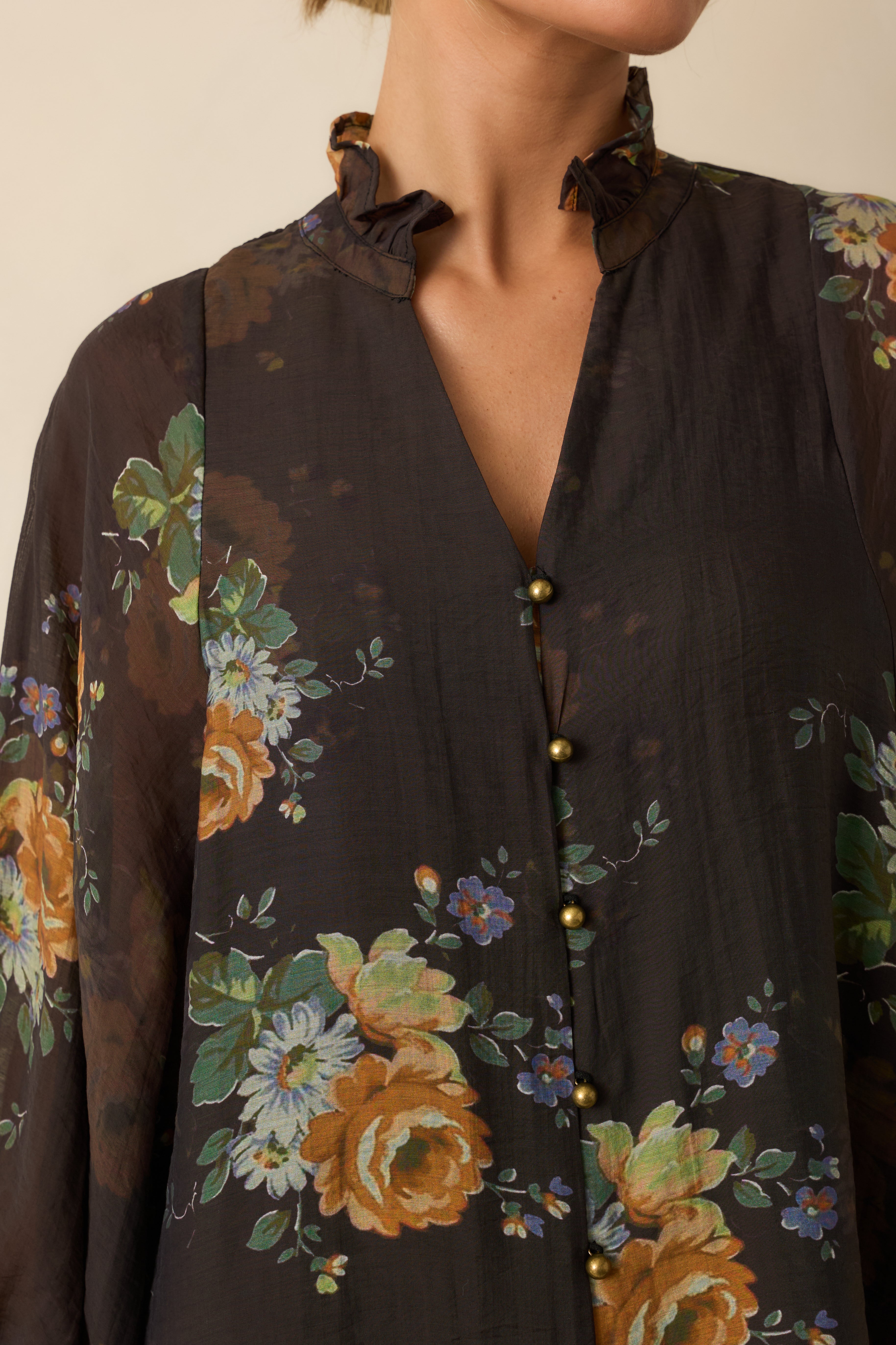Close-up of the ruffled neckline, gold buttons, and floral print detailing on the brown blouse.