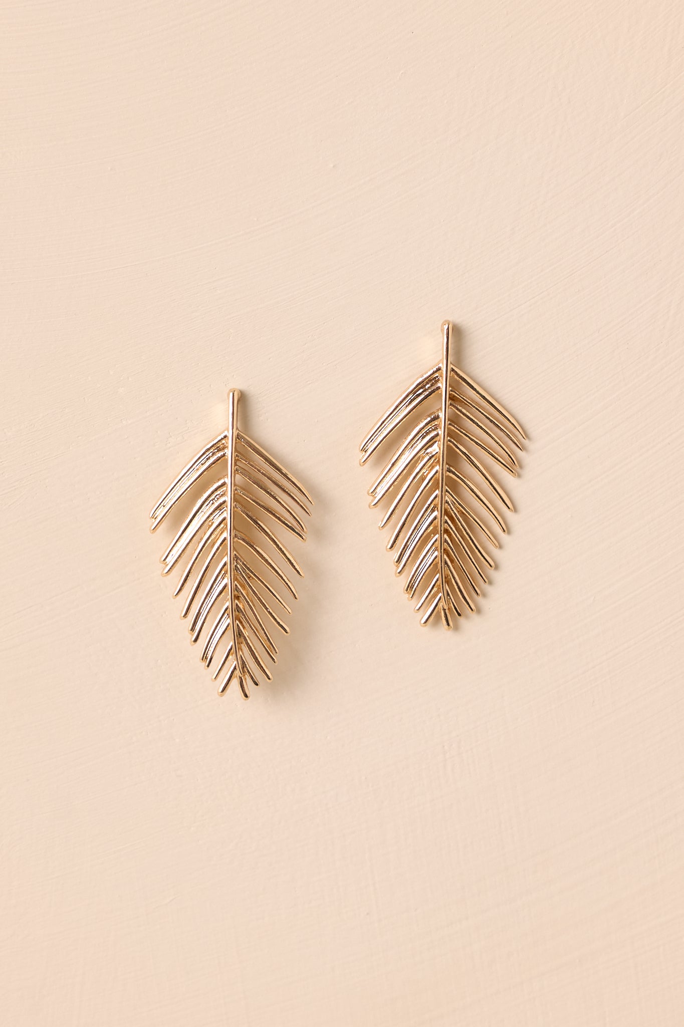A close-up photo of the palm leaf-shaped gold earrings with a smooth finish and ridged details shown against a beige background.