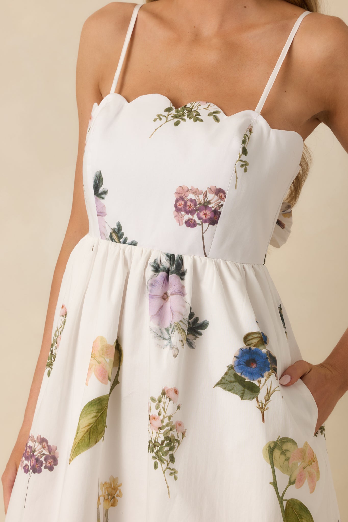 Close-up of white midi dress showing scalloped neckline and hem, floral print, and tie-back detail on fitted bodice.
