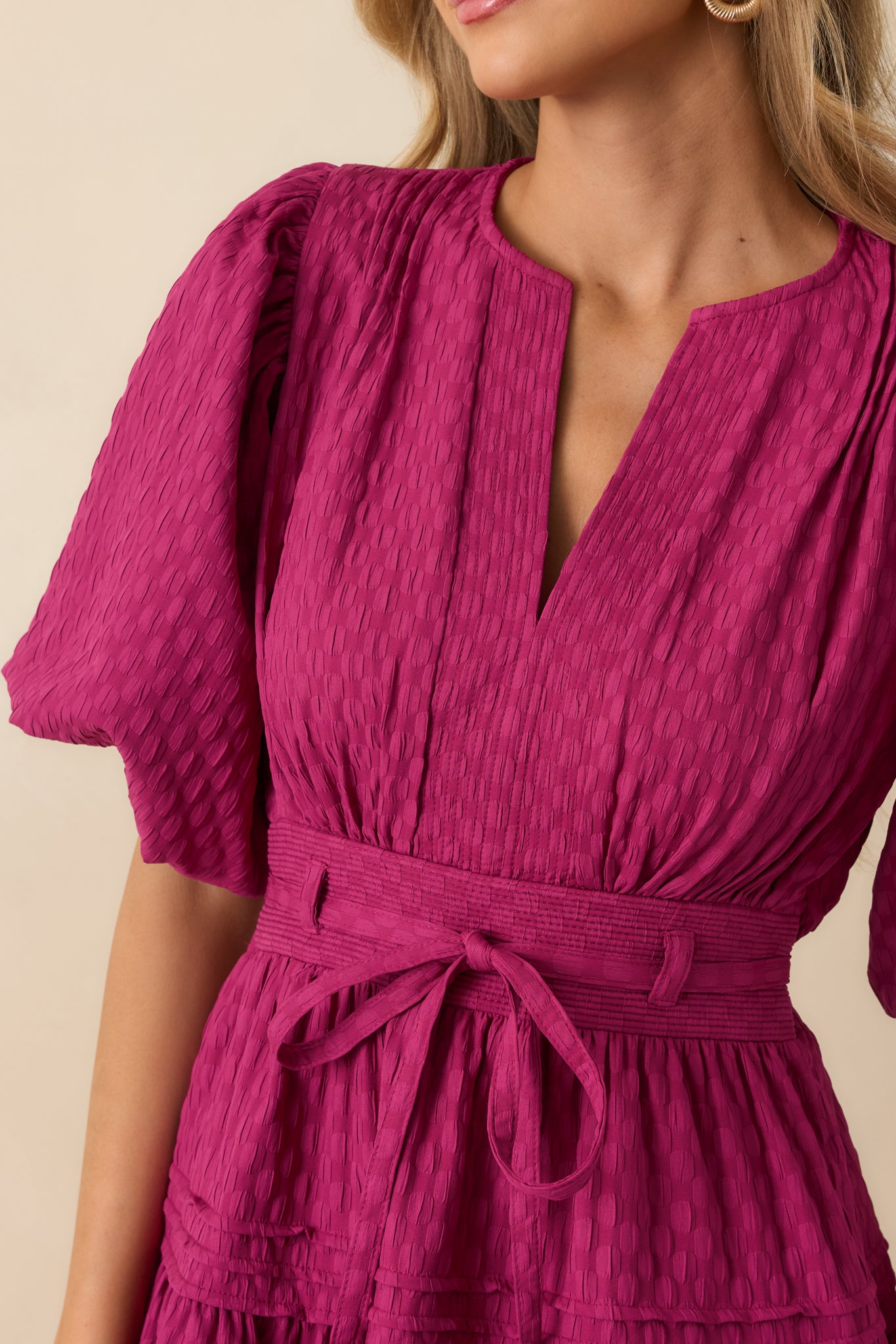 A close view reveals the textured woven fabric, pleated bodice, and adjustable tie waist with precise stitching.