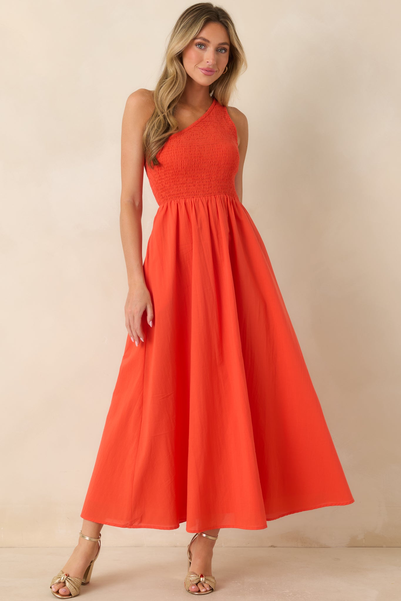 Coral-orange cotton maxi dress featuring a one-shoulder neckline, fully smocked bodice, and a flowing A-line skirt with hidden side pockets.