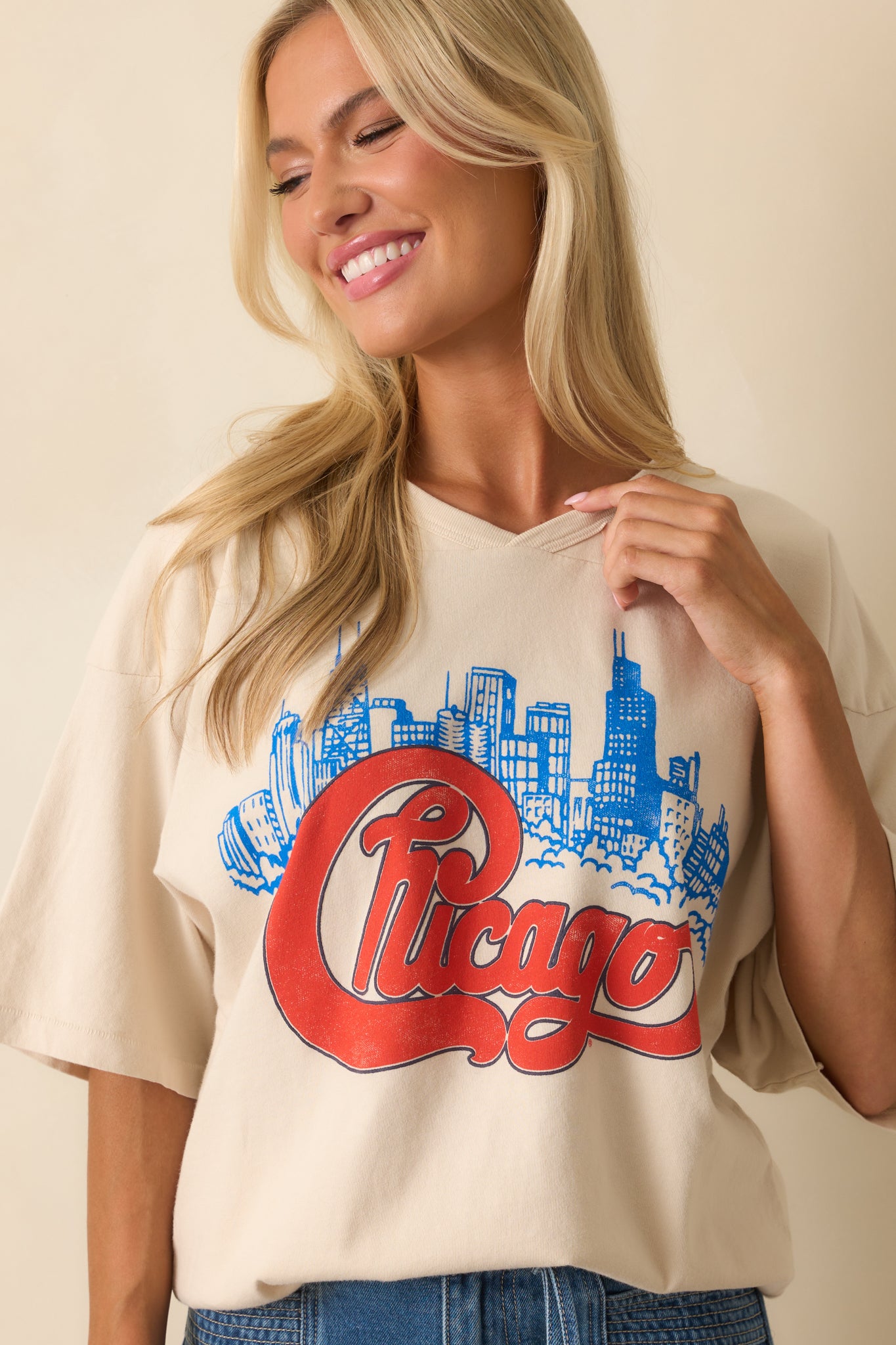 Cropped view of ivory oversized T-shirt highlighting bold “Chicago” script in red over detailed blue skyline graphic.