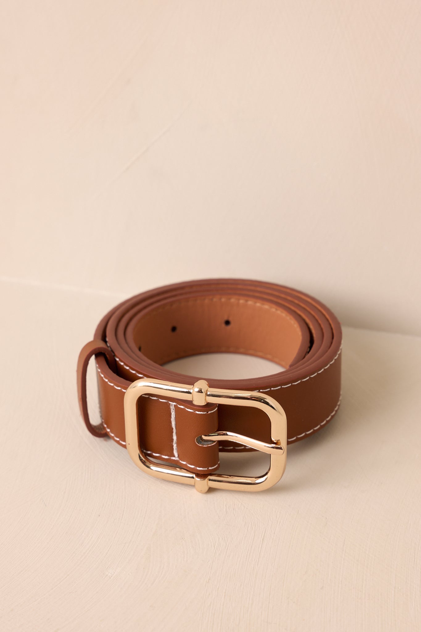 Cropped view of a brown faux leather belt with ivory stitched edges and a gold square pin buckle.