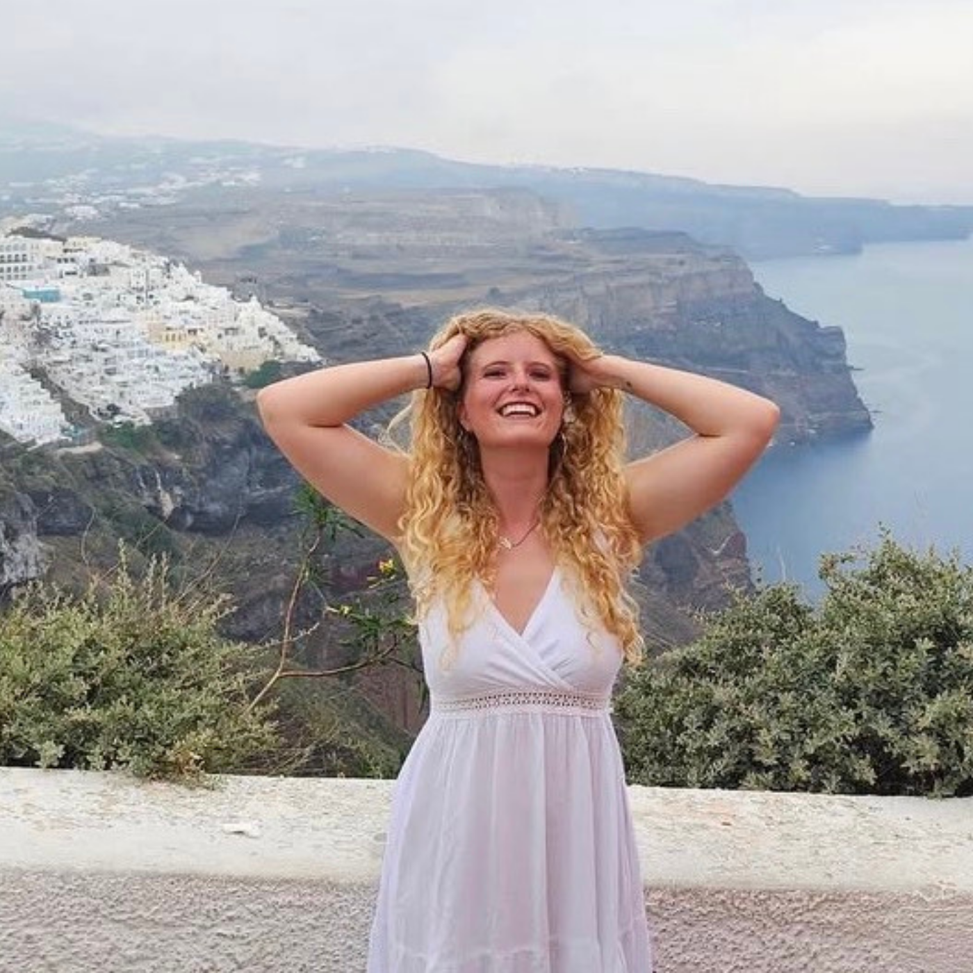 Woman in a white dress with a scenic background of a coastal town on a cliff, over the ocean.