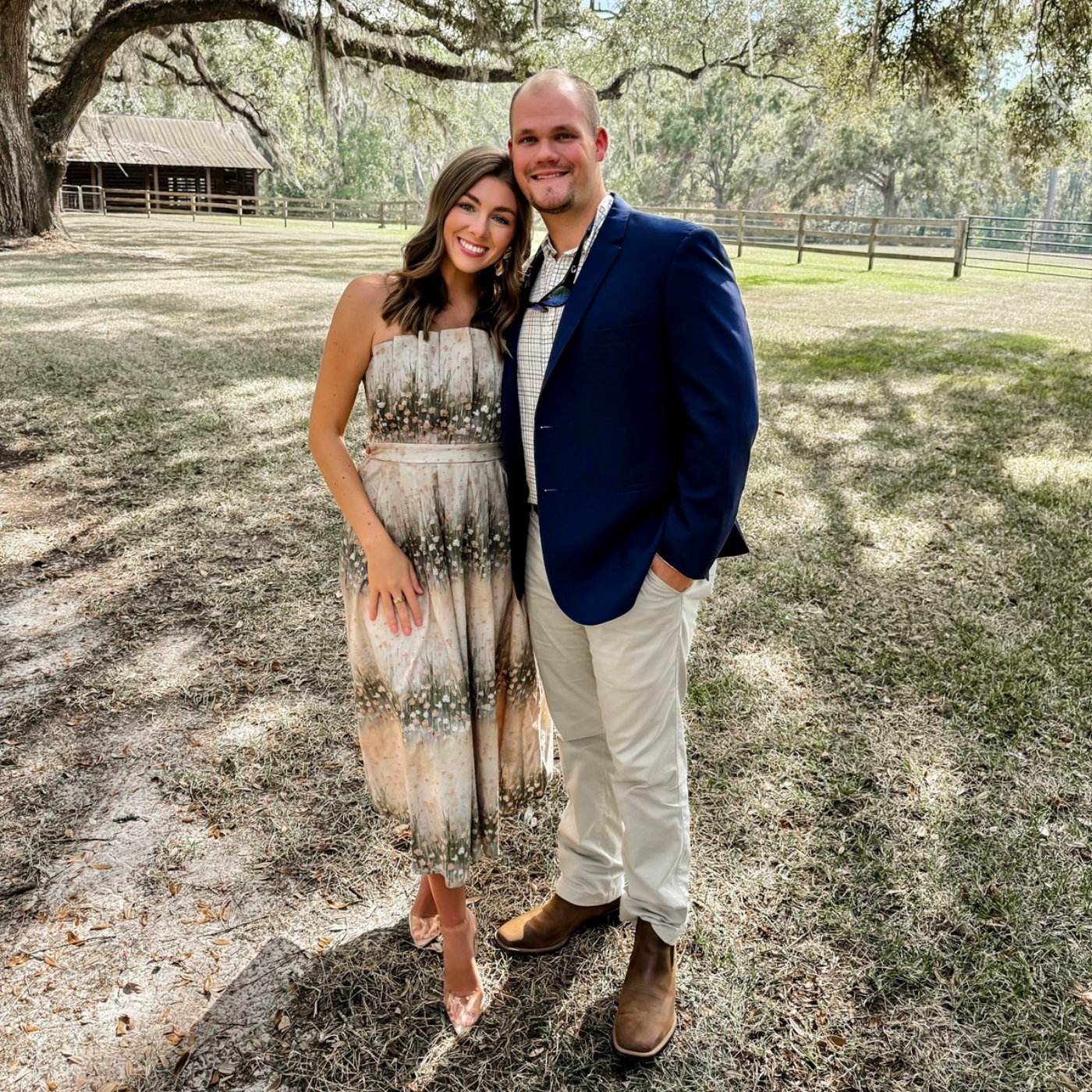 Man and woman standing in a field with trees and a wooden structure in the background
