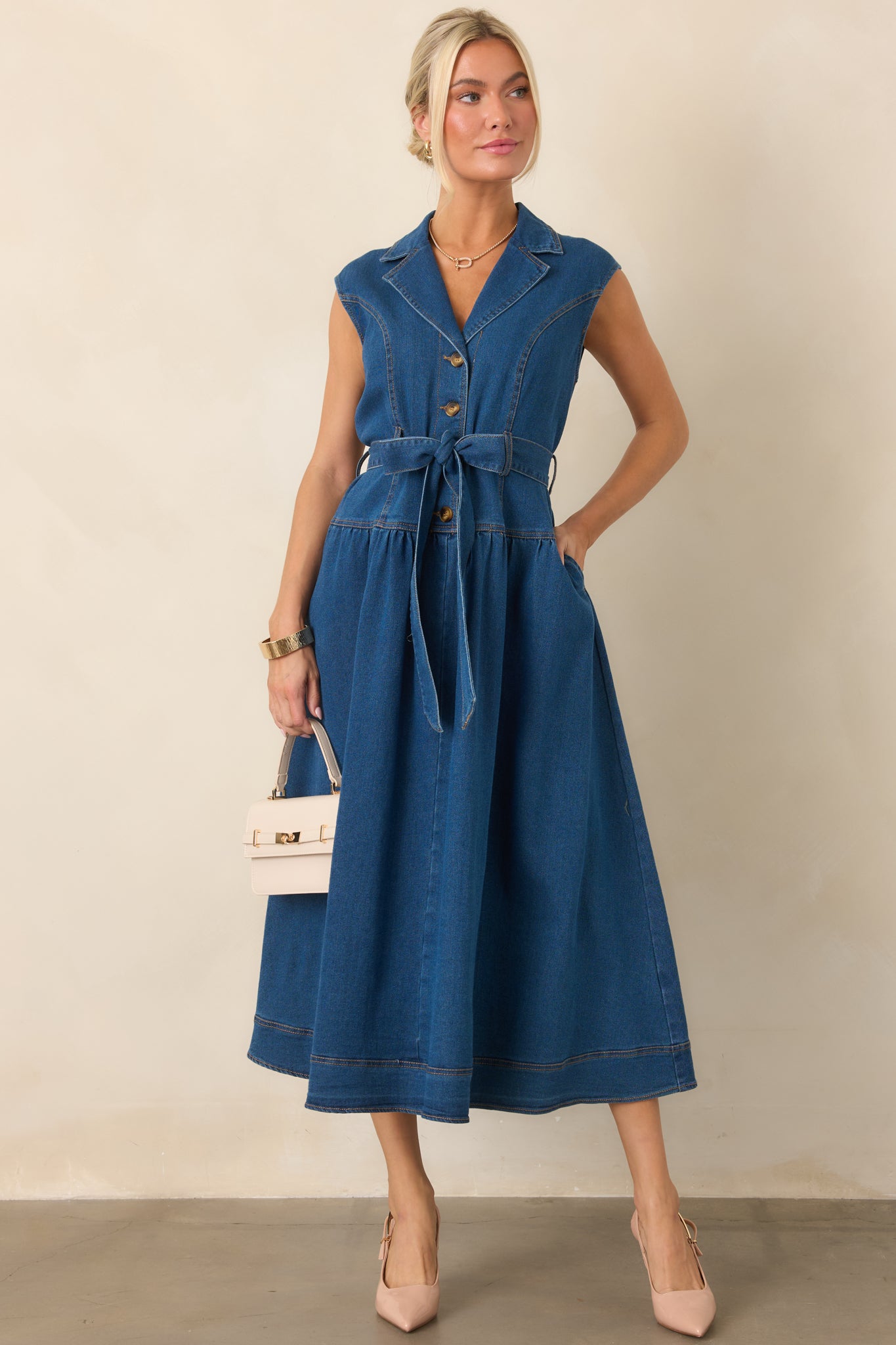 Dark wash denim midi dress featuring a sleeveless silhouette, tailored collared neckline, gold-tone front buttons, removable self-tie belt, and side pockets.