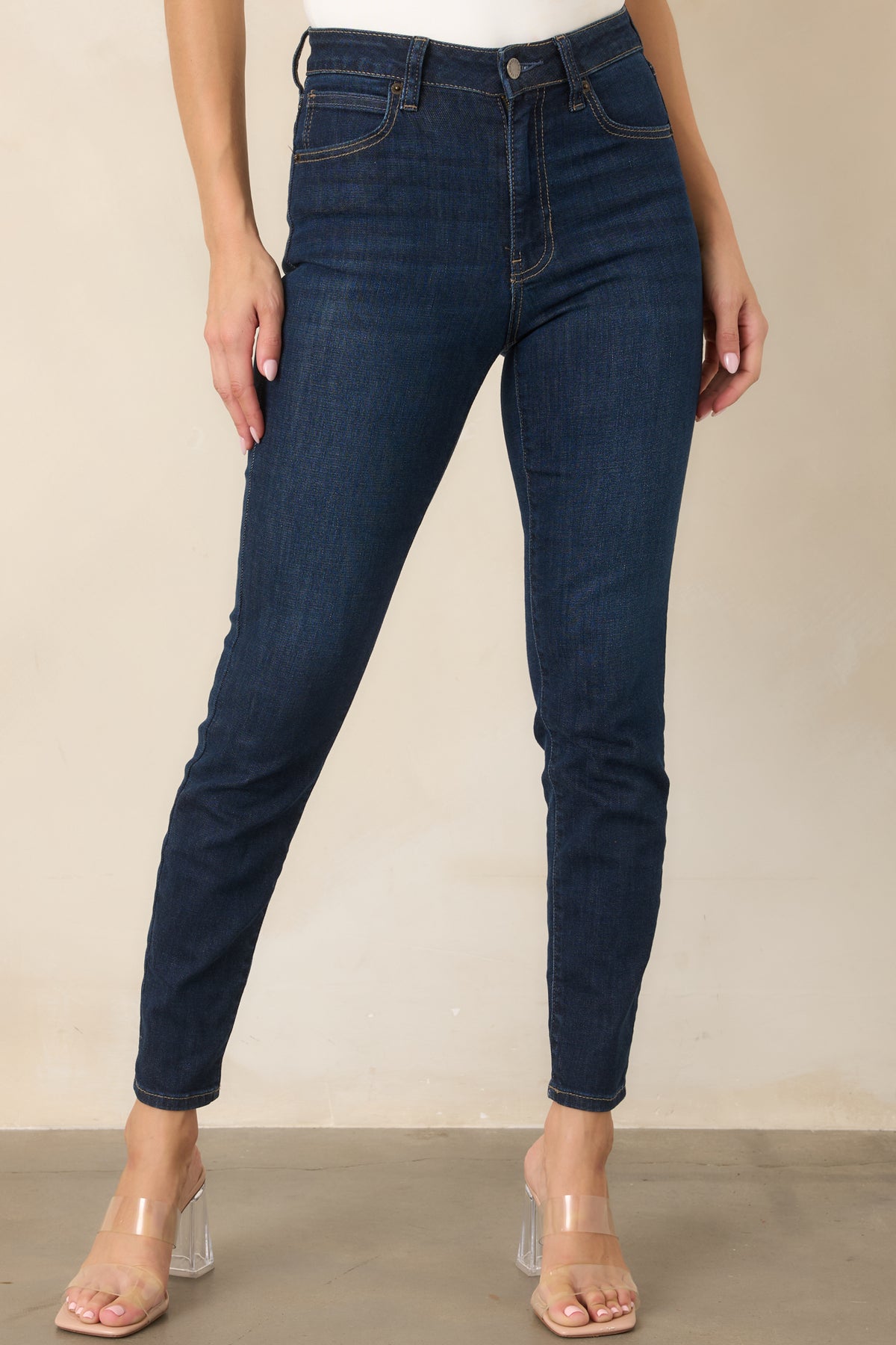Dark wash jeans shown in detail with a functional front pocket, belt loop, zipper closure, and neatly finished tan stitching.