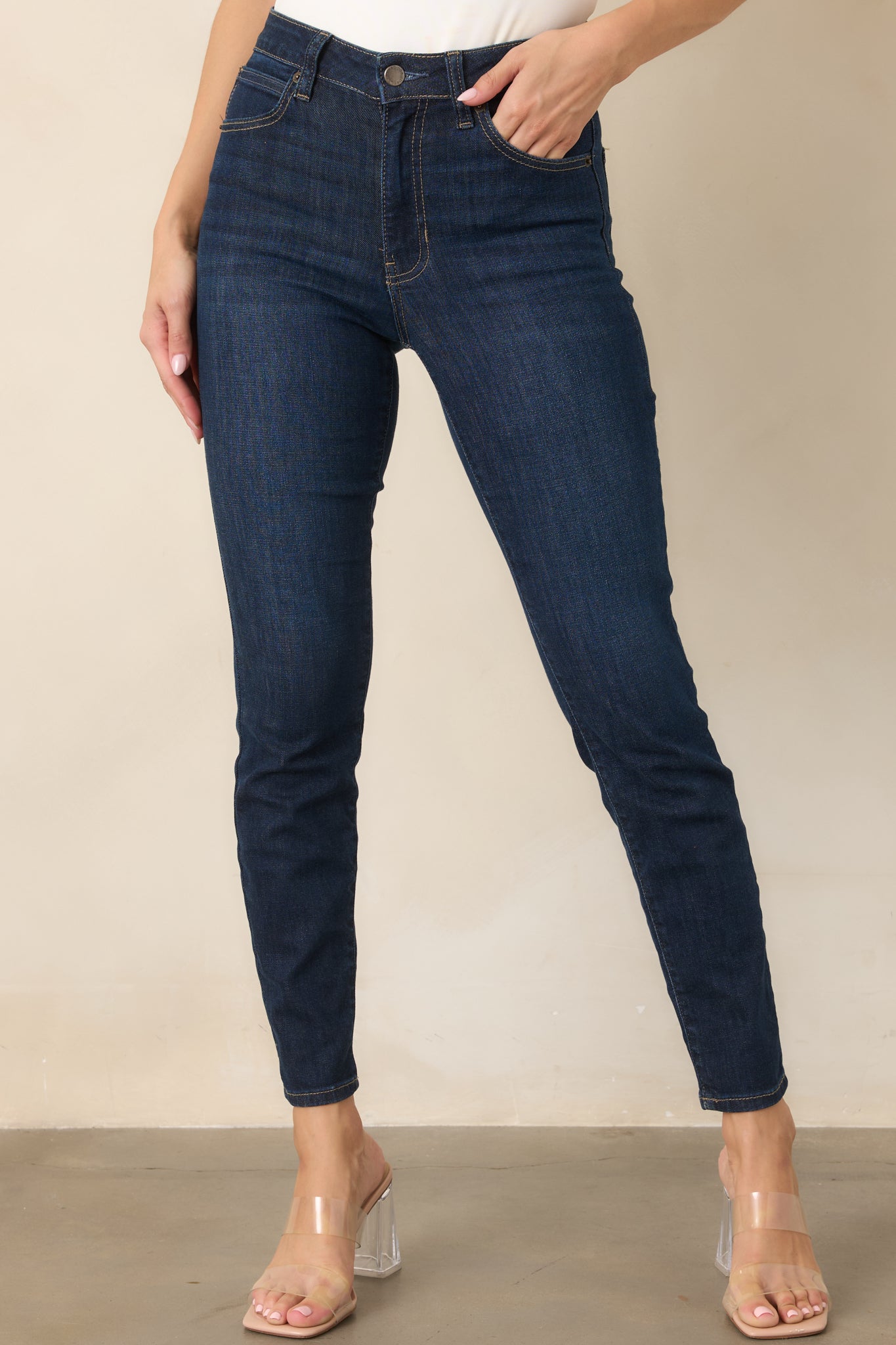 Close view of the dark wash jeans highlighting the high-rise waistband, belt loops, zipper fly, and button closure with tan stitching.