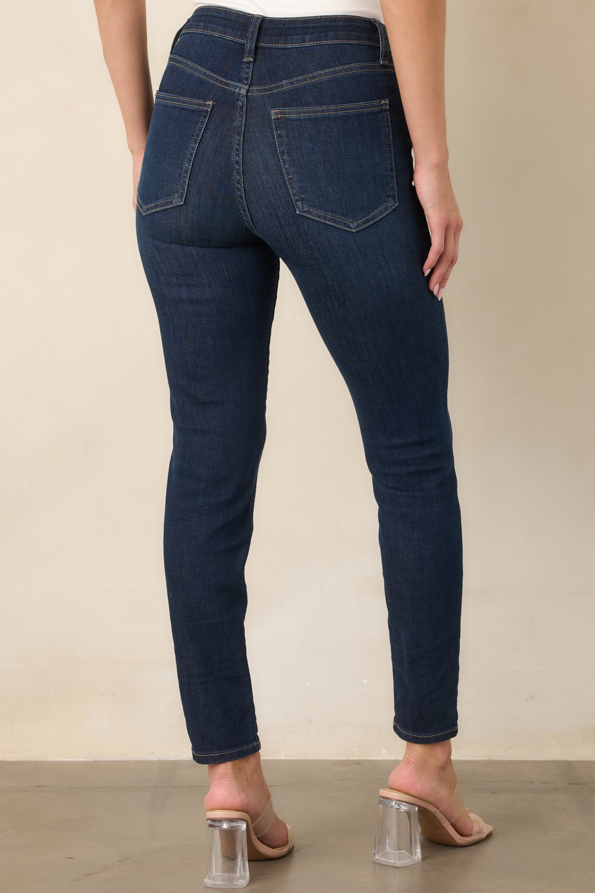 Rear view of the dark wash skinny jeans showcasing the high-rise waistband, functional back pockets, fitted legs, and tan topstitching throughout.