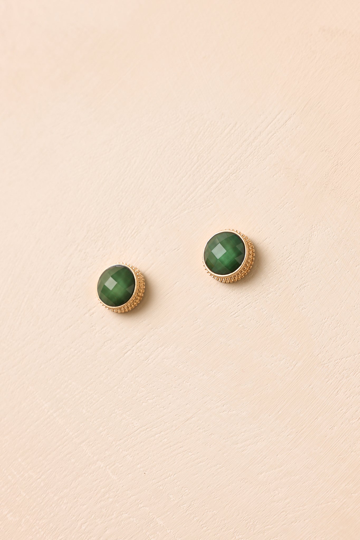 Deep green faceted stud earrings displayed against a beige background featuring a polished gold halo frame and timeless round silhouette.