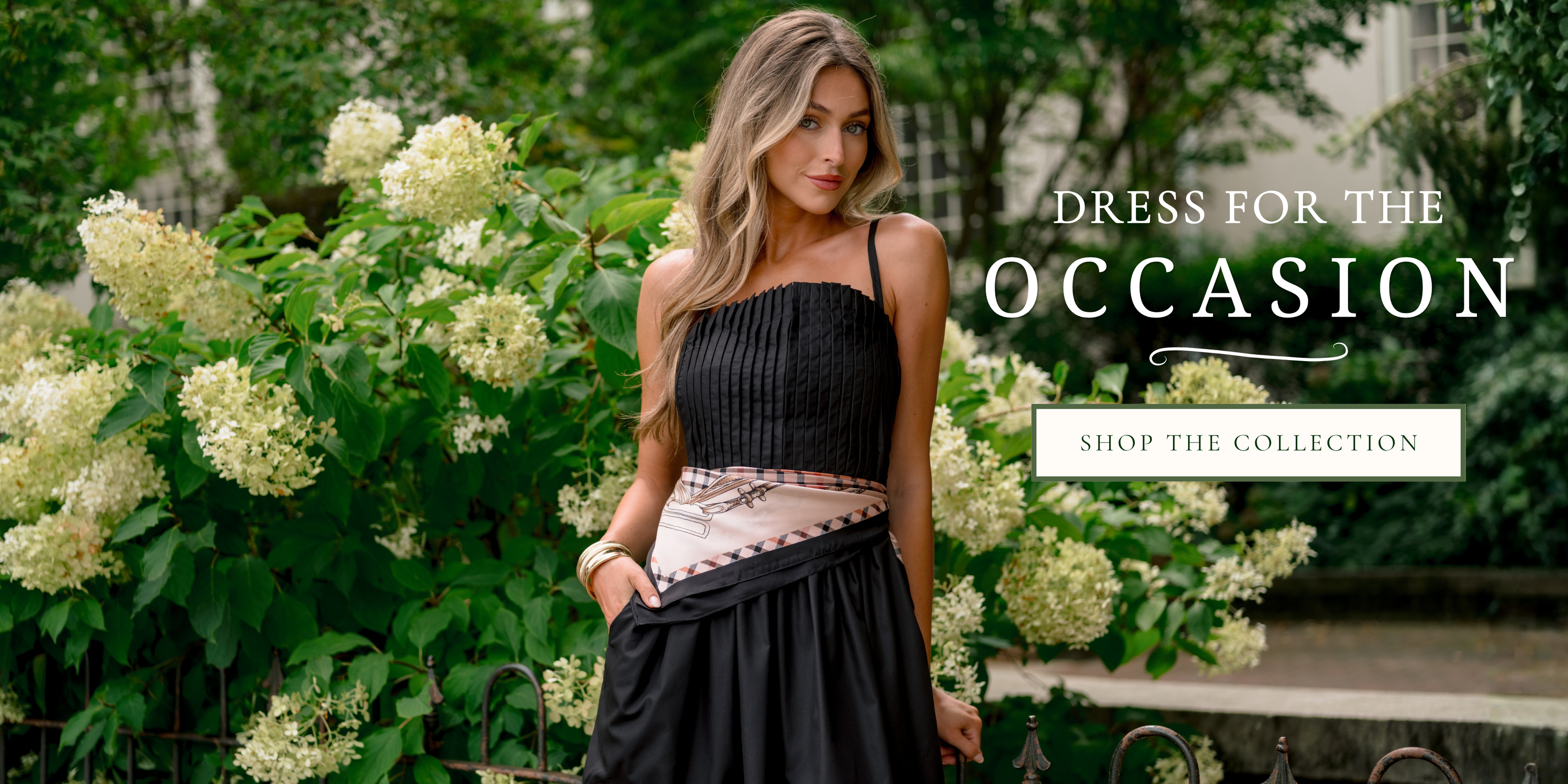 Model wearing a black pleated midi dress with a printed waist scarf, standing in a garden with white hydrangeas. Text reads, "Dress for the Occasion" with a "Shop the Collection" button.