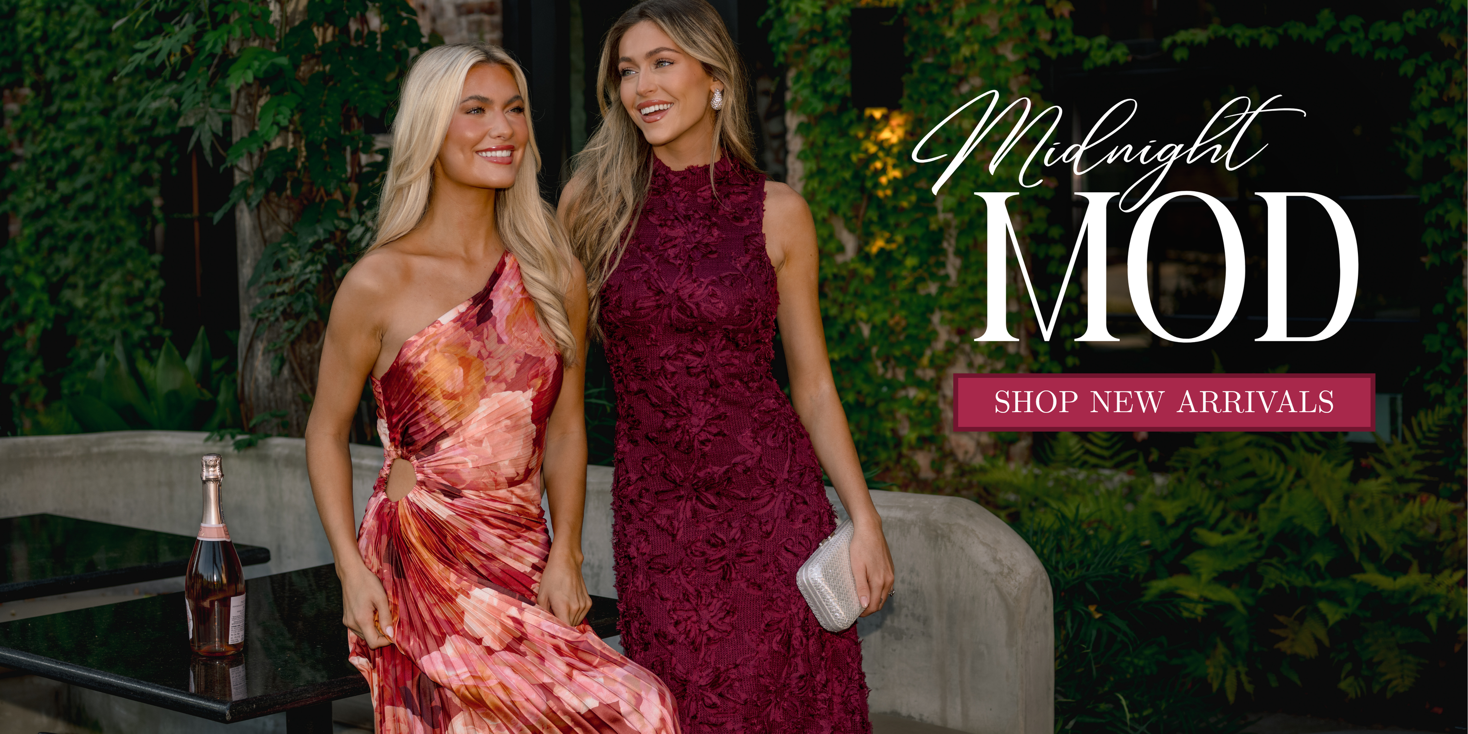 Two women wearing elegant evening dresses, one in a floral one-shoulder dress and the other in a burgundy textured dress, standing outdoors at night. Text reads “Midnight Mod” and “Shop New Arrivals.”