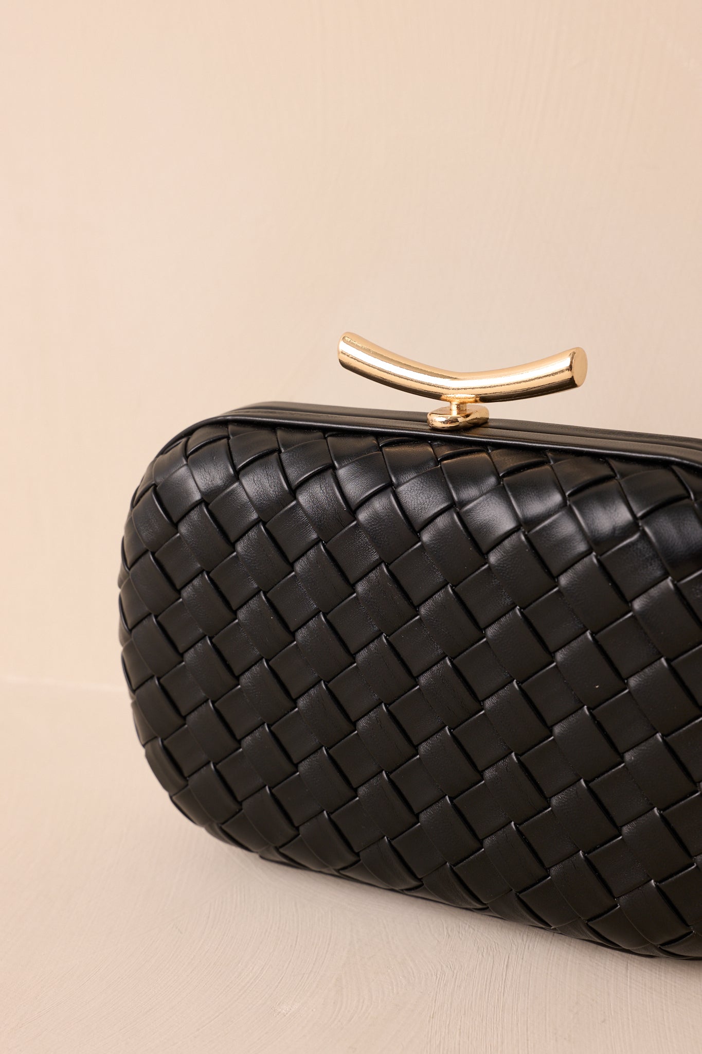 A close-up detail of the black clutch showing the woven exterior, polished clasp, and structured hard-shell body.