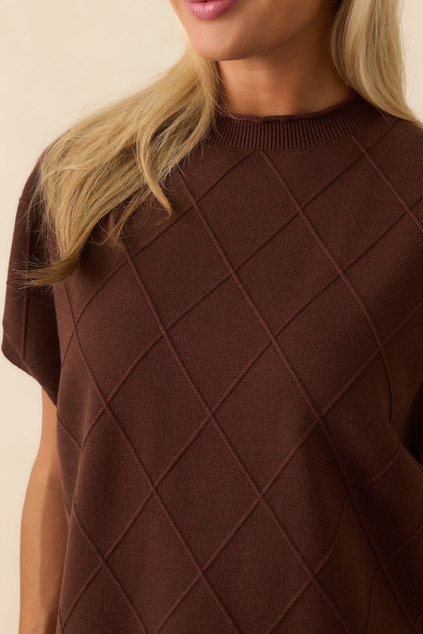 Detailed shot of brown sweater showing argyle stitch pattern, rolled crew neckline, and folded sleeve cuffs.