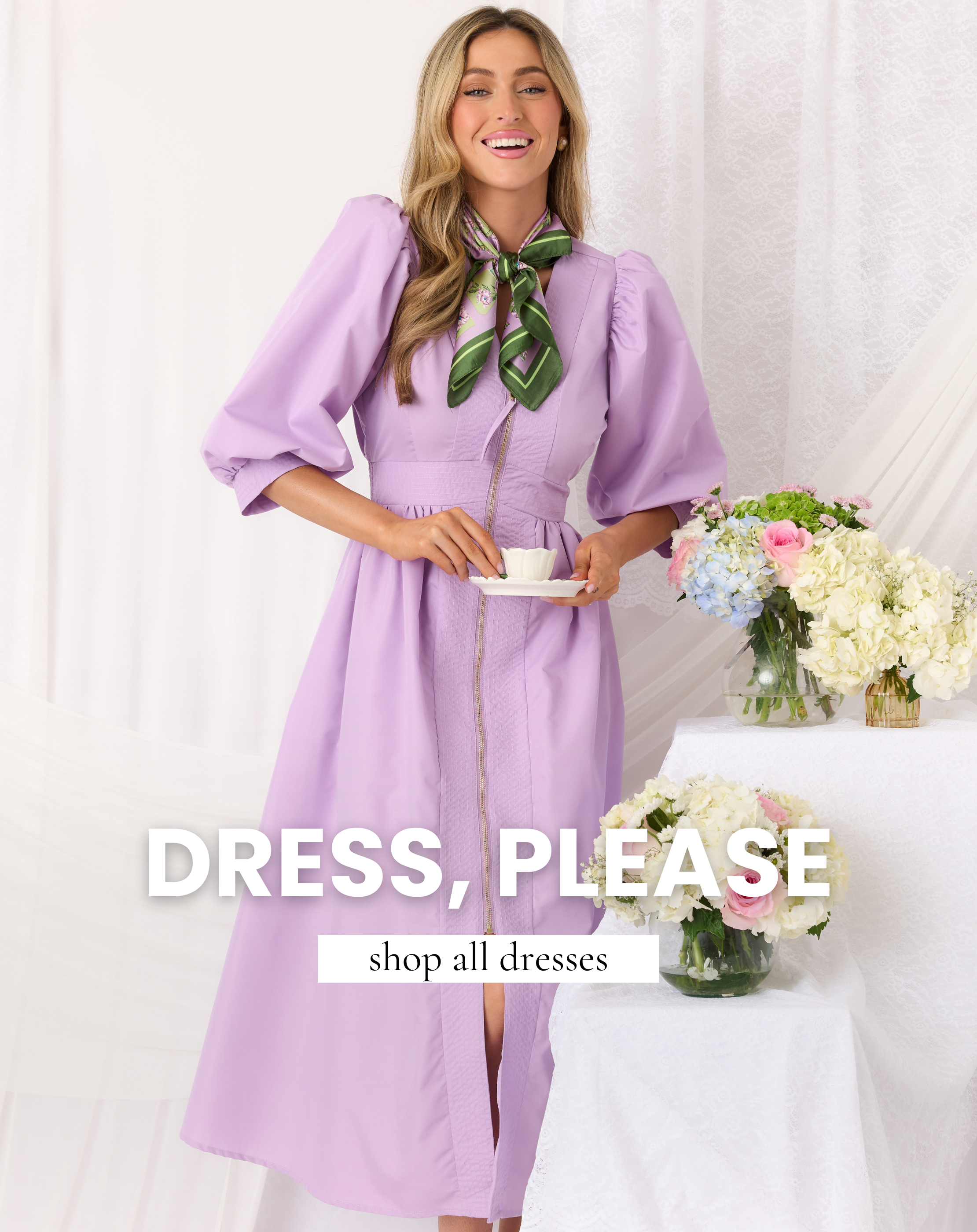 Model wearing a lavender puff sleeve midi dress with a green neck scarf, standing beside pastel floral arrangements and holding a teacup. This dress is featured un our All Dresses collection