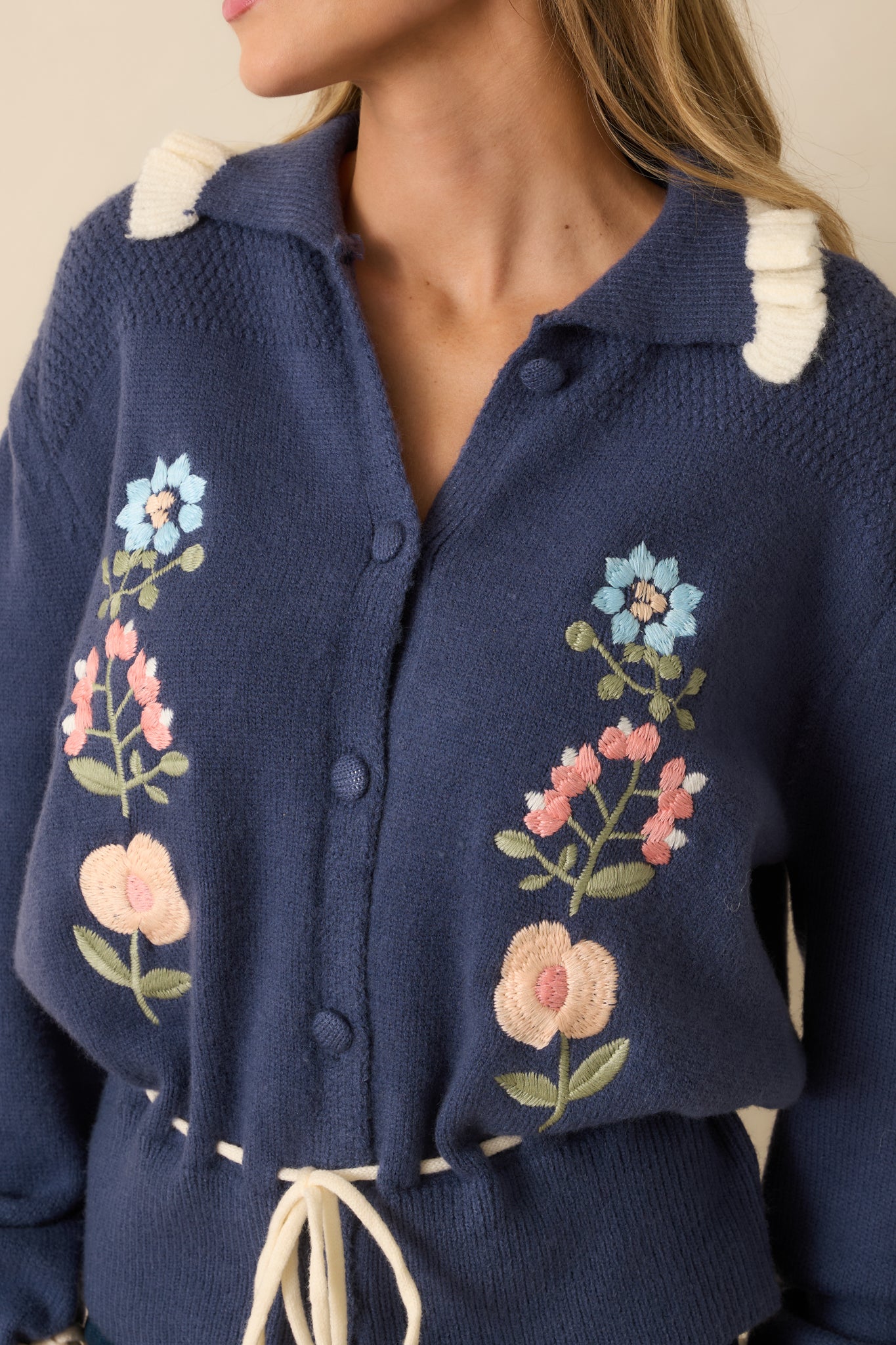 Tight detail showing the colorful floral embroidery, ivory collar ruffle, and tie cuffs on the dusty blue cardigan.