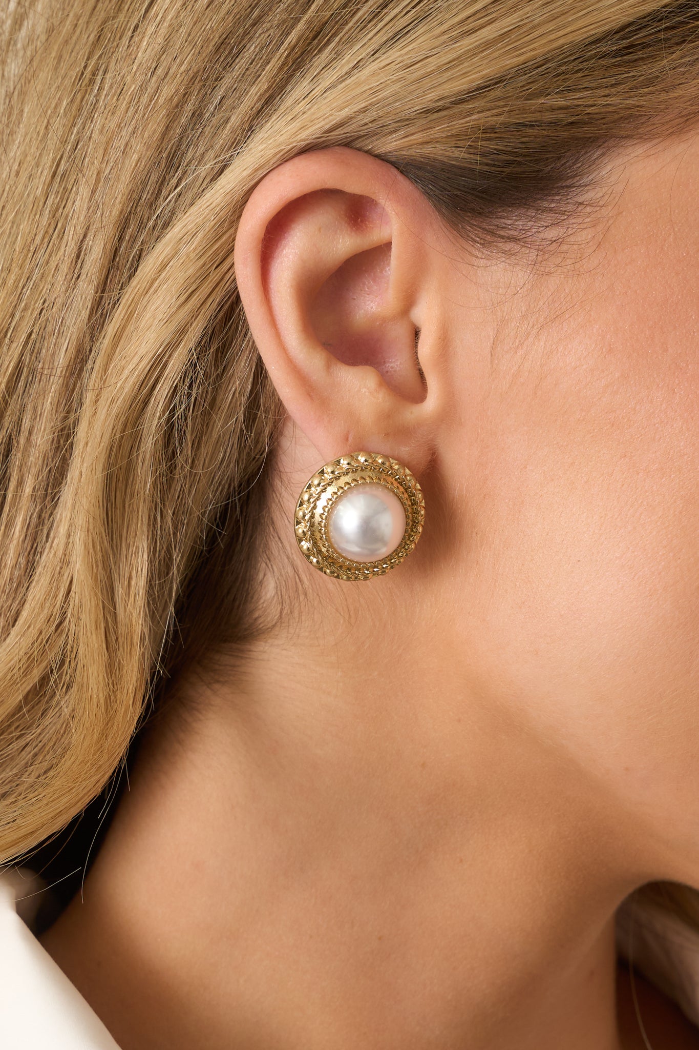 A close-up of one earring showing the central ivory pearl, thick textured gold border, braided edge, and secure post backing.