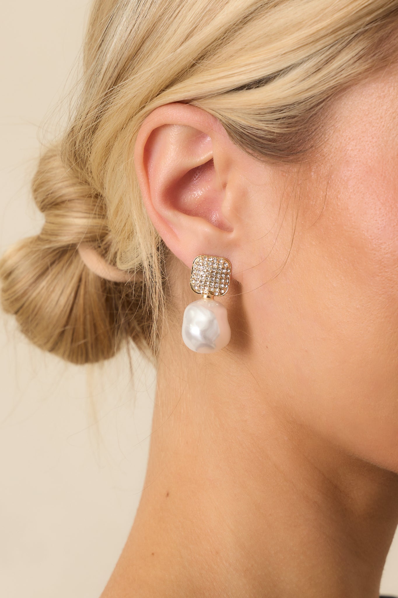 A cropped view of the earrings showing square gold studs with rhinestones and dangling ivory faux baroque pearls.
