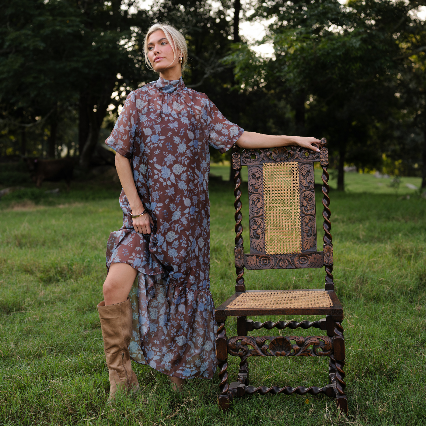 Woman in a blue and brown floral, sheer dress with a high neckline and a brown slip underneath. She is standing in a field of tall grass with an antique wooden chair.