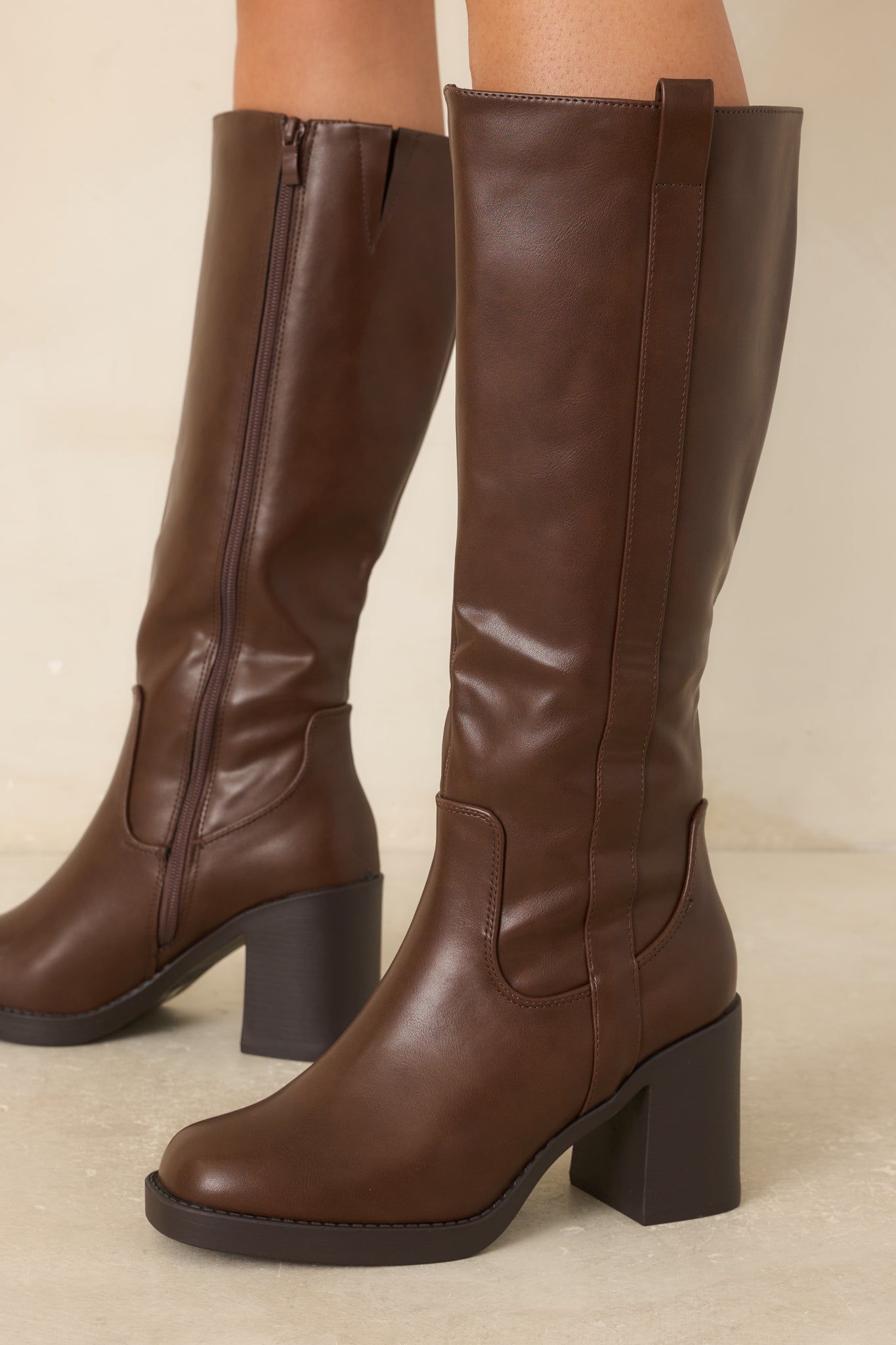 Tall faux leather boots featuring a softly squared toe, sturdy block heel, slim platform sole, vertical outer shaft strap, and structured silhouette against a beige background.