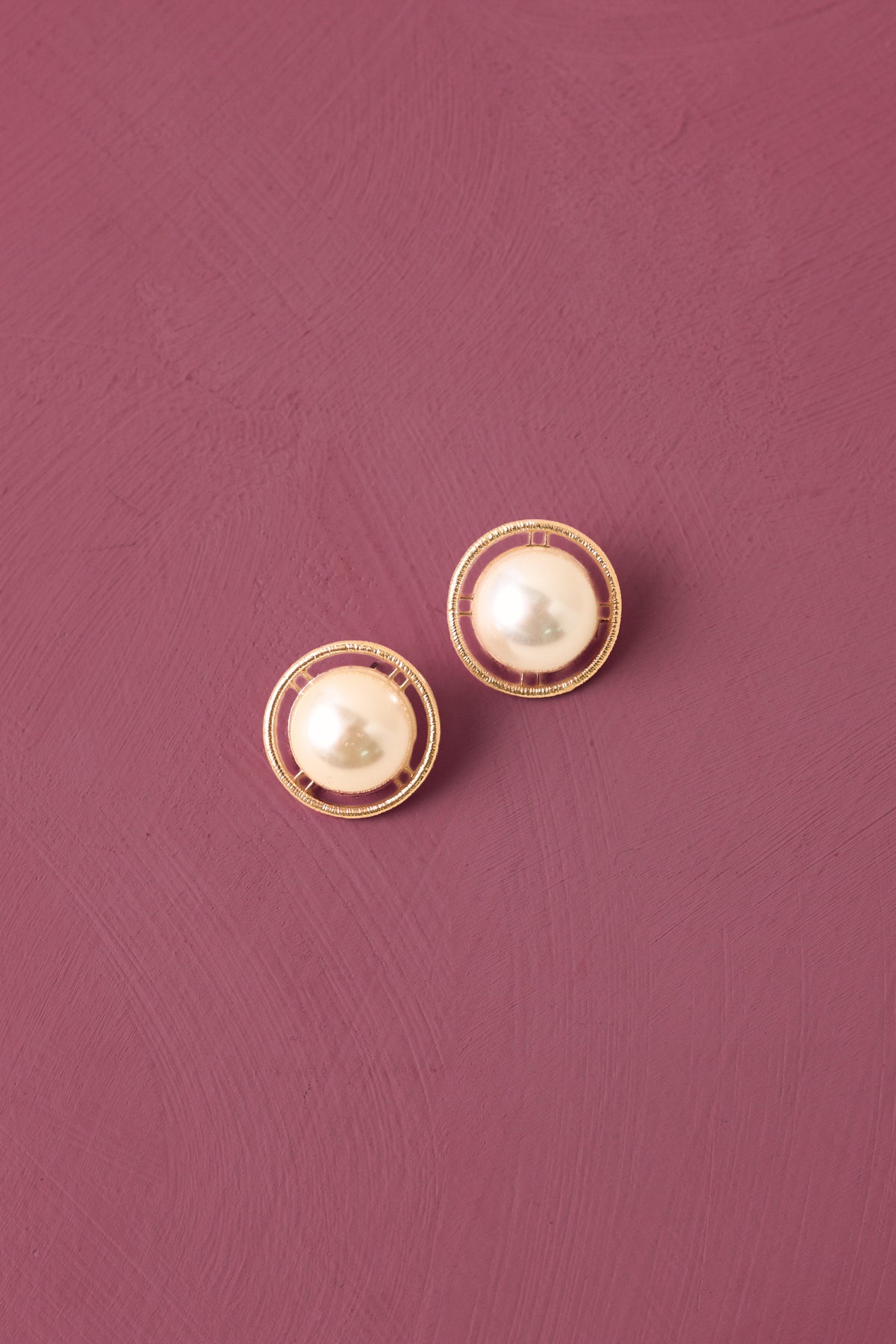 Pearl studs with a gold hardware frame against a deep mauve backdrop. 