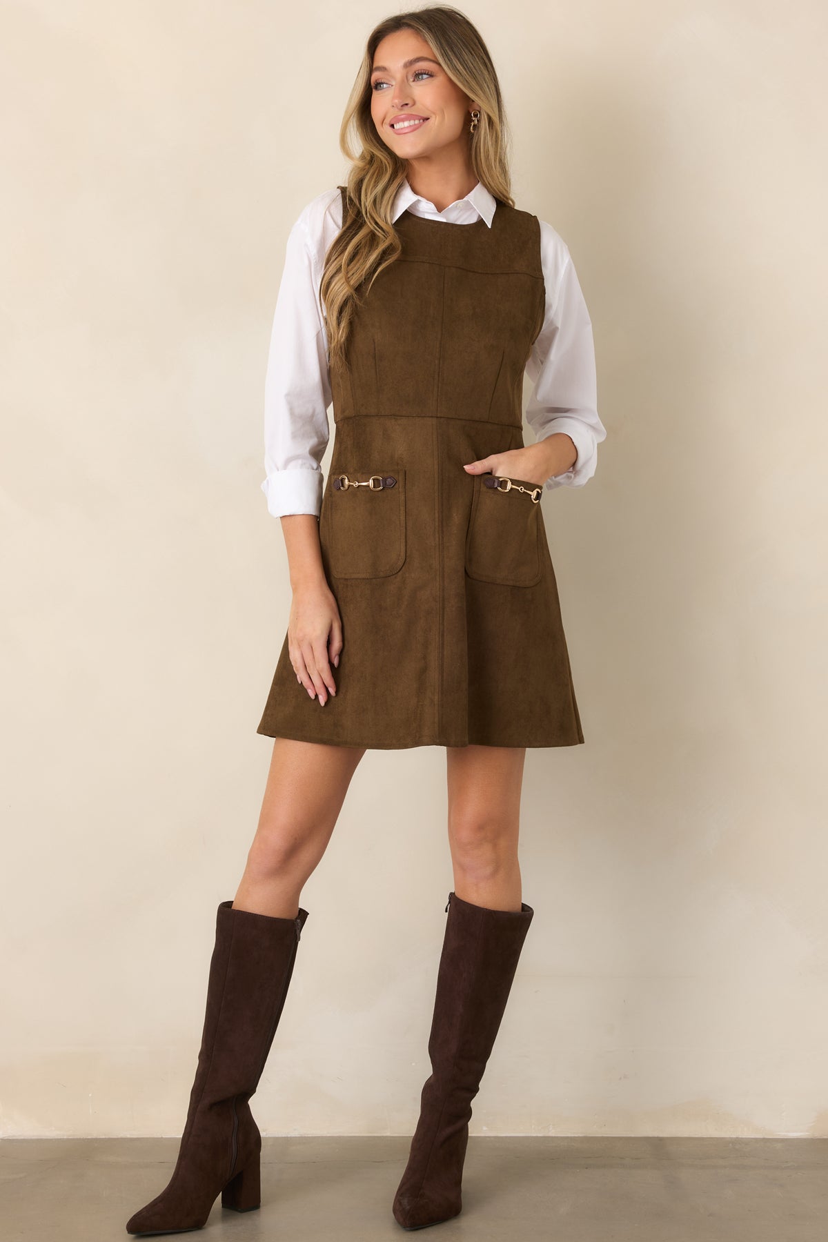 Sleeveless faux suede mini dress with prominent seam detailing, a defined waist, flared skirt, and front patch pockets accented by gold-tone hardware.