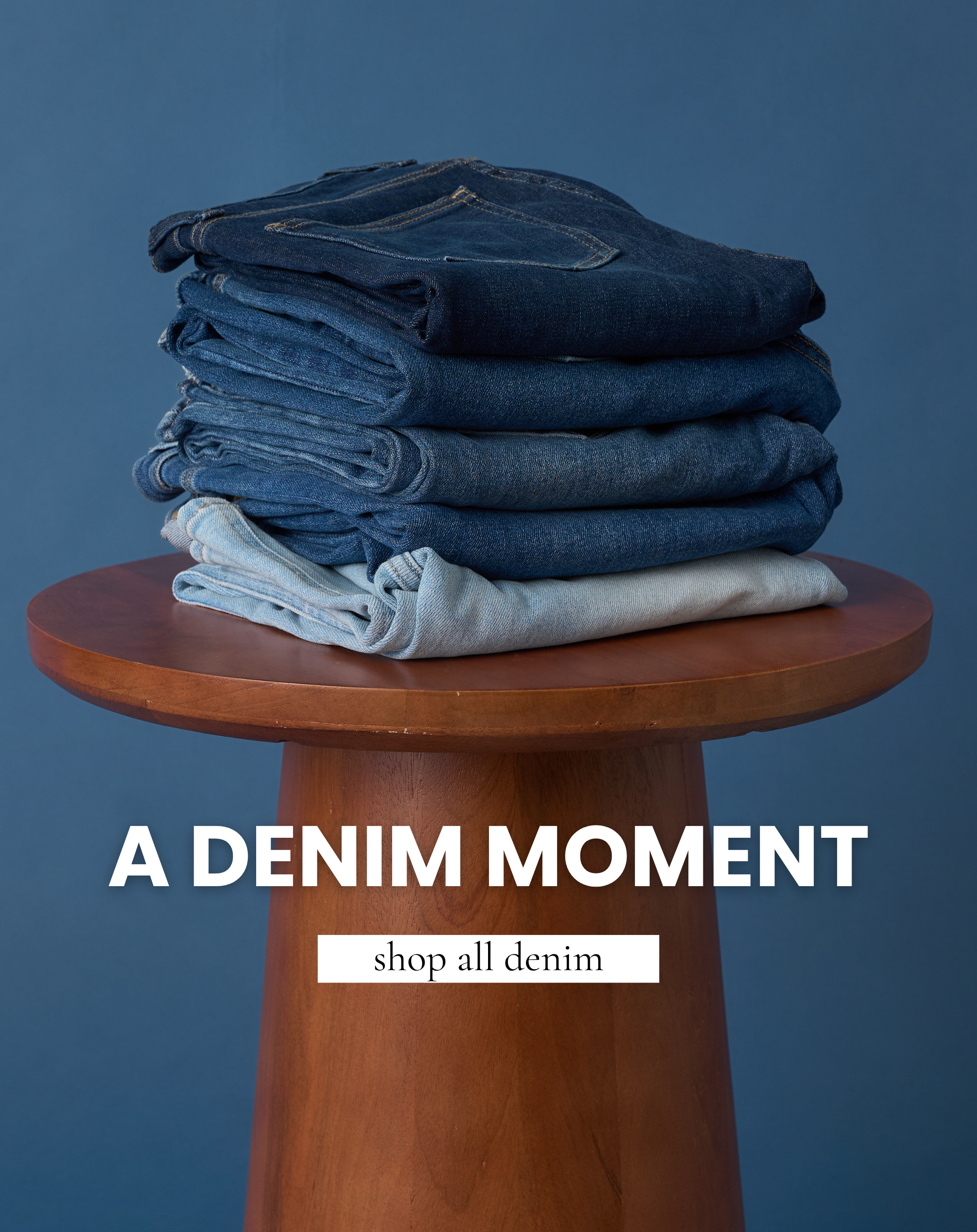 Stack of folded denim jeans in various shades of blue displayed on a wooden table against a blue background. Text reads “A Denim Moment” and “Shop All Denim.”