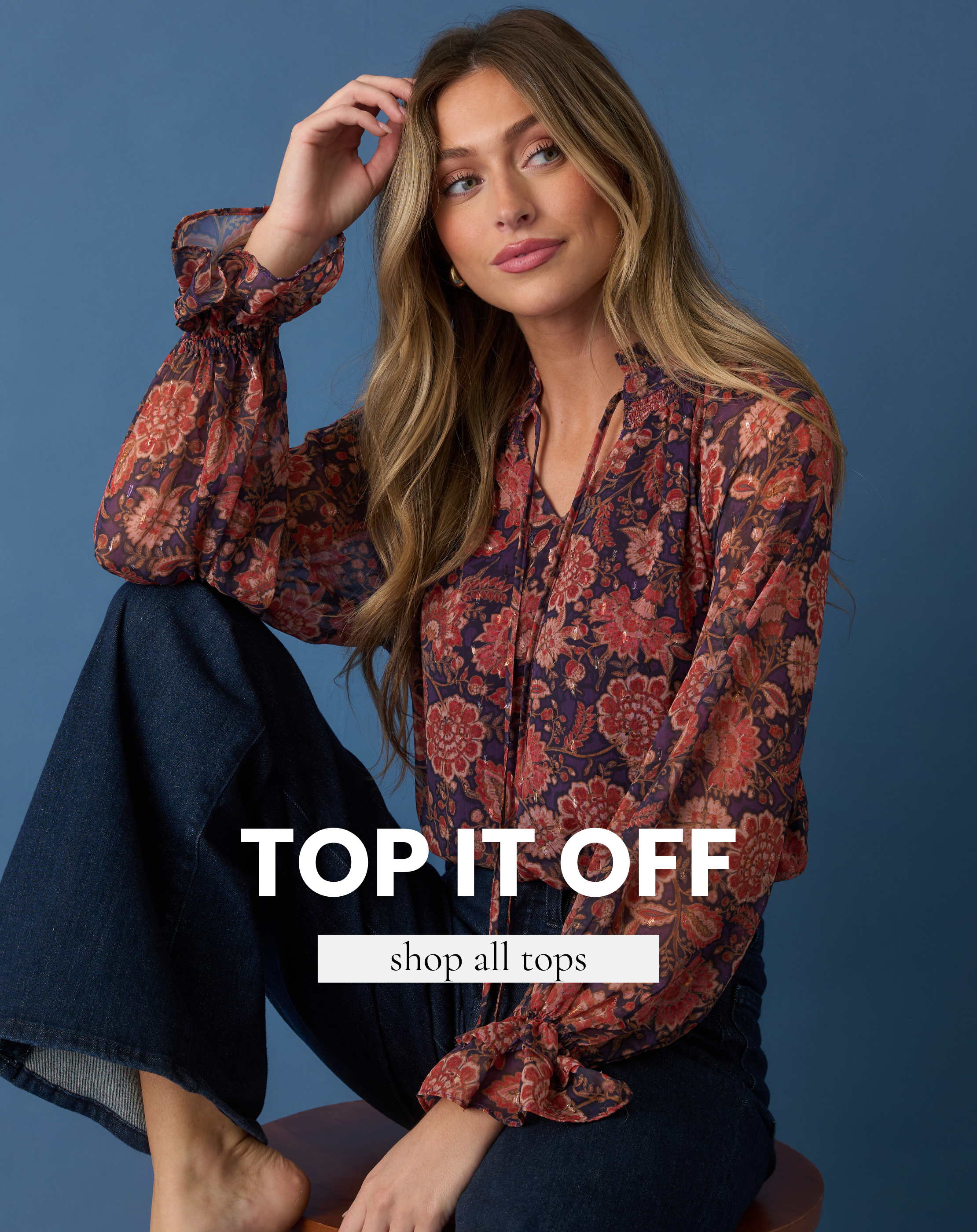 Model wearing a floral long-sleeve blouse with dark denim jeans against a blue background. Text reads “Top It Off” and “Shop All Tops.”