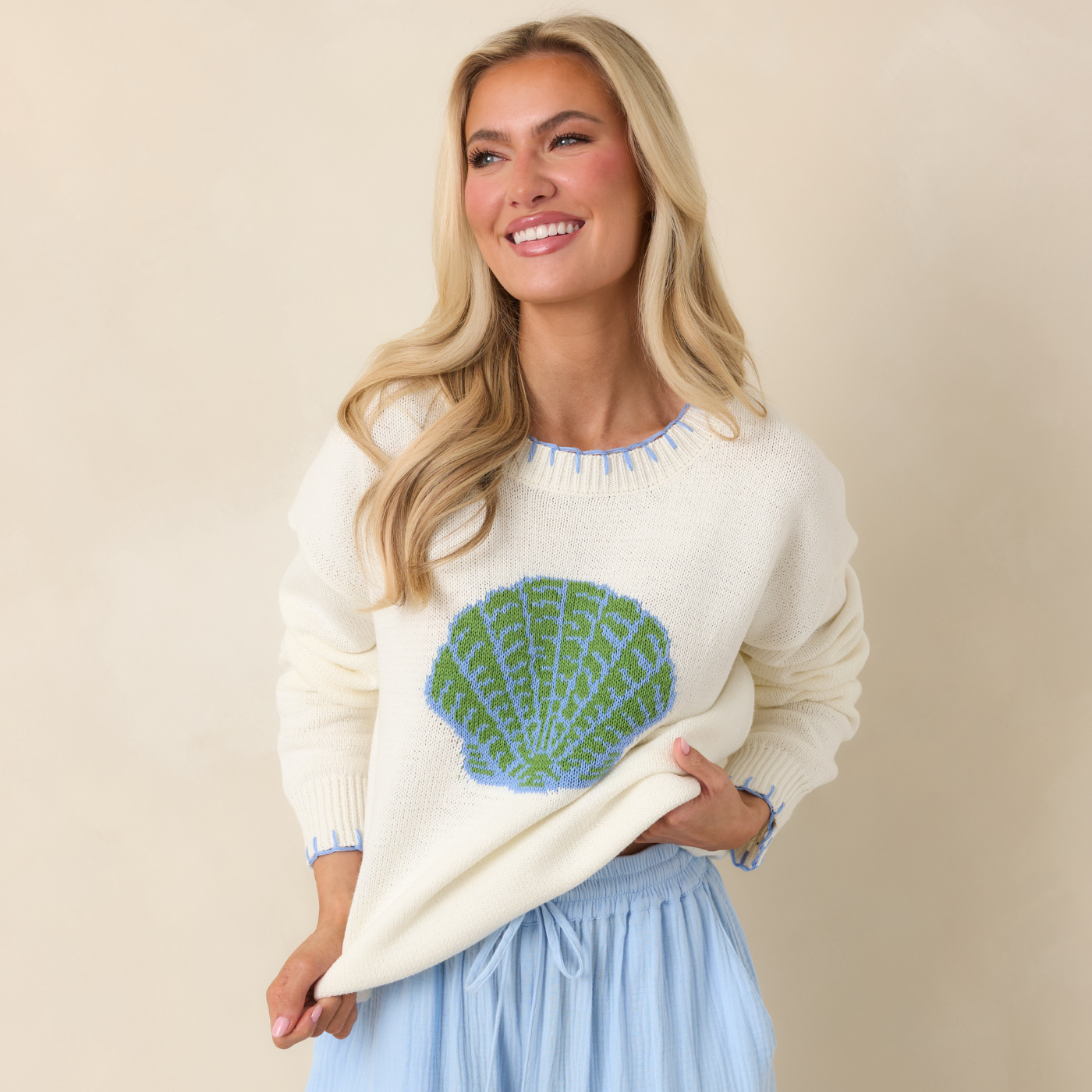 Woman wearing a cream sweater with a green shell design on a beige background.