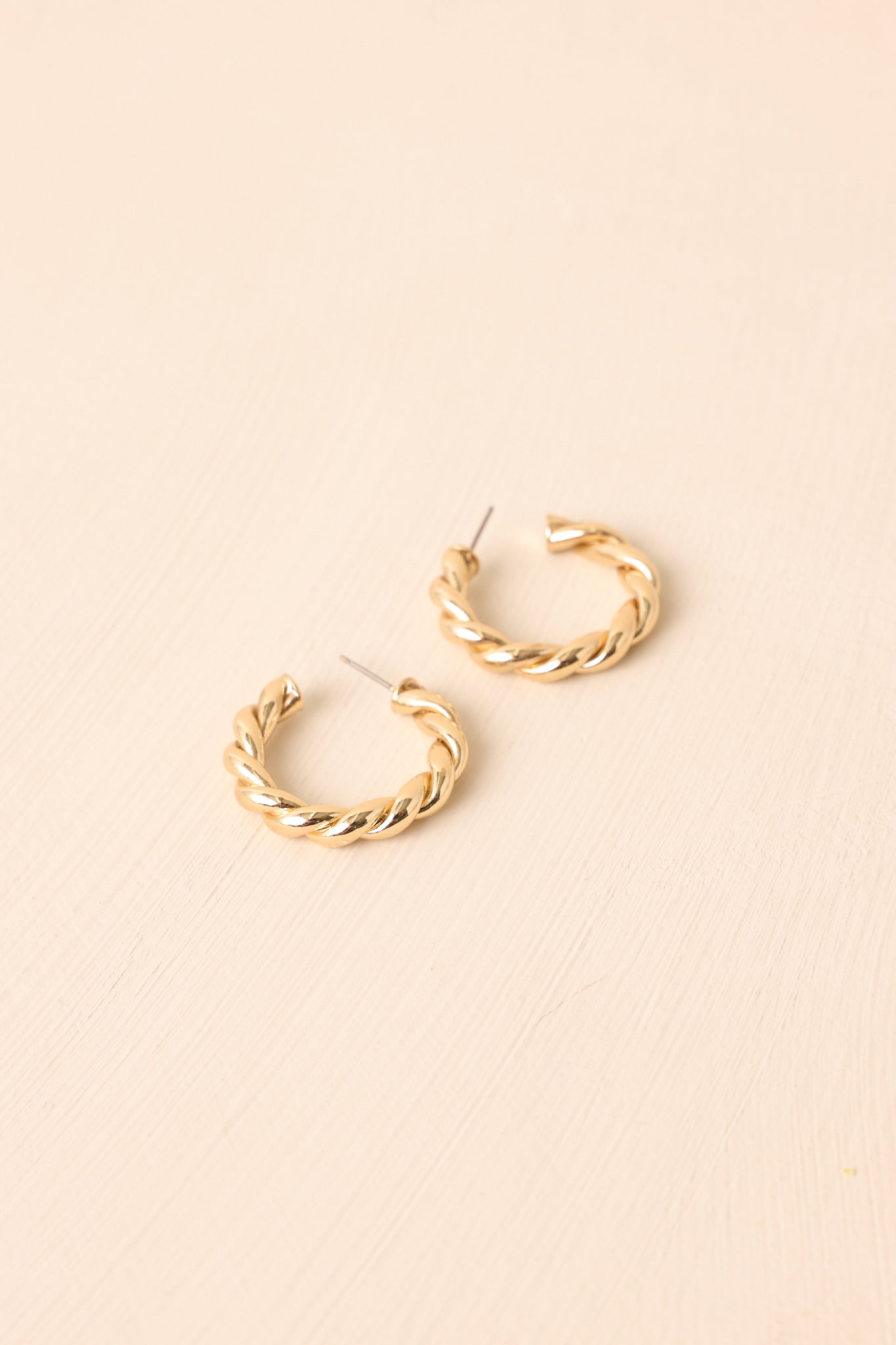 Flat lay of the earrings on a beige surface, showcasing the reflective gold finish and elegant twist in the open design.