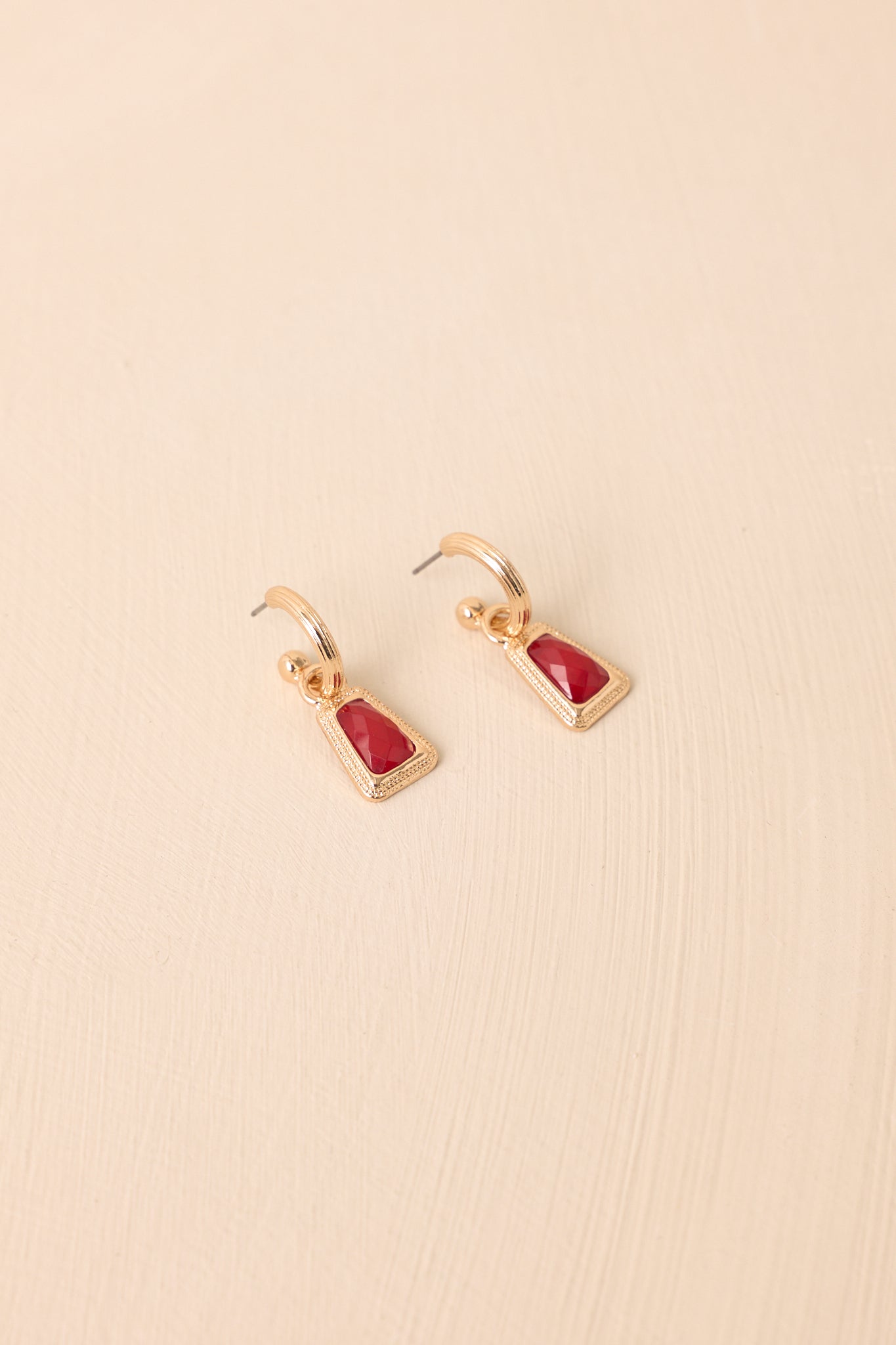 Flat lay of the earrings on a beige surface highlighting the red stone centerpiece and the huggie-style clasp.