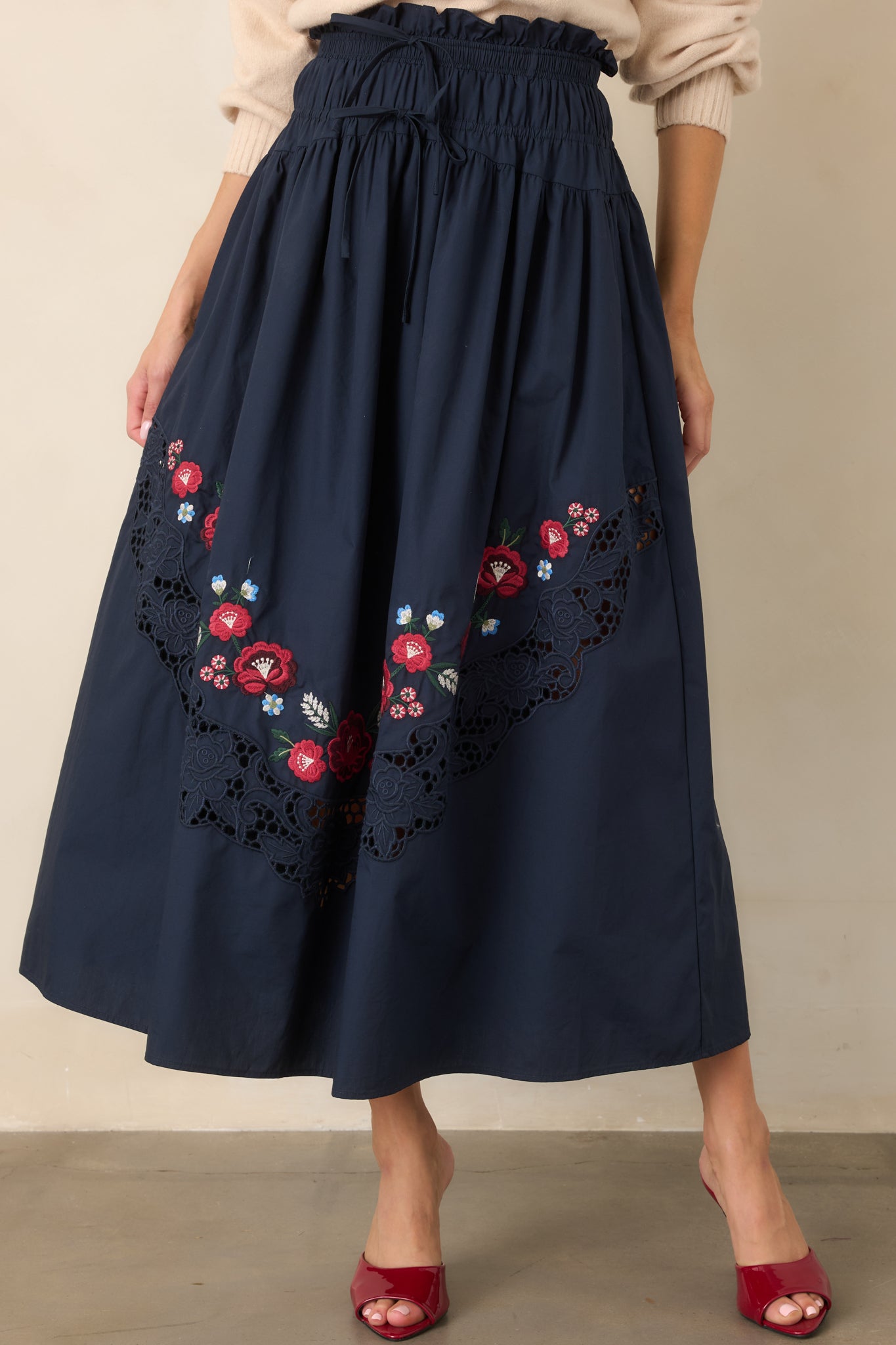 Full, flowing cotton skirt featuring colorful red, blue, green, and ivory floral embroidery, a wide openwork lace inset, and an elastic gathered waistband with decorative front ties.