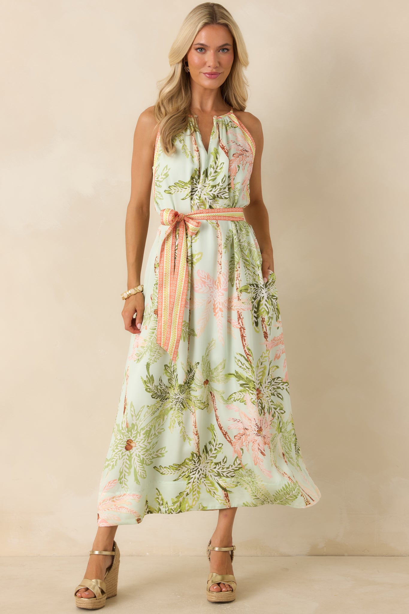 Flowing maxi dress featuring a sleeveless neckline with gathered detailing, a defined waist sash, and a watercolor palm print in green, blush, and soft neutral tones.
