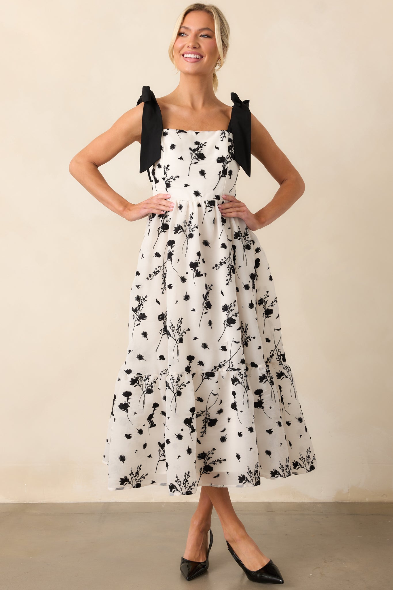 A flowy ivory midi dress with black floral embroidery, featuring adjustable straps and a hidden back zipper for a polished, feminine look.