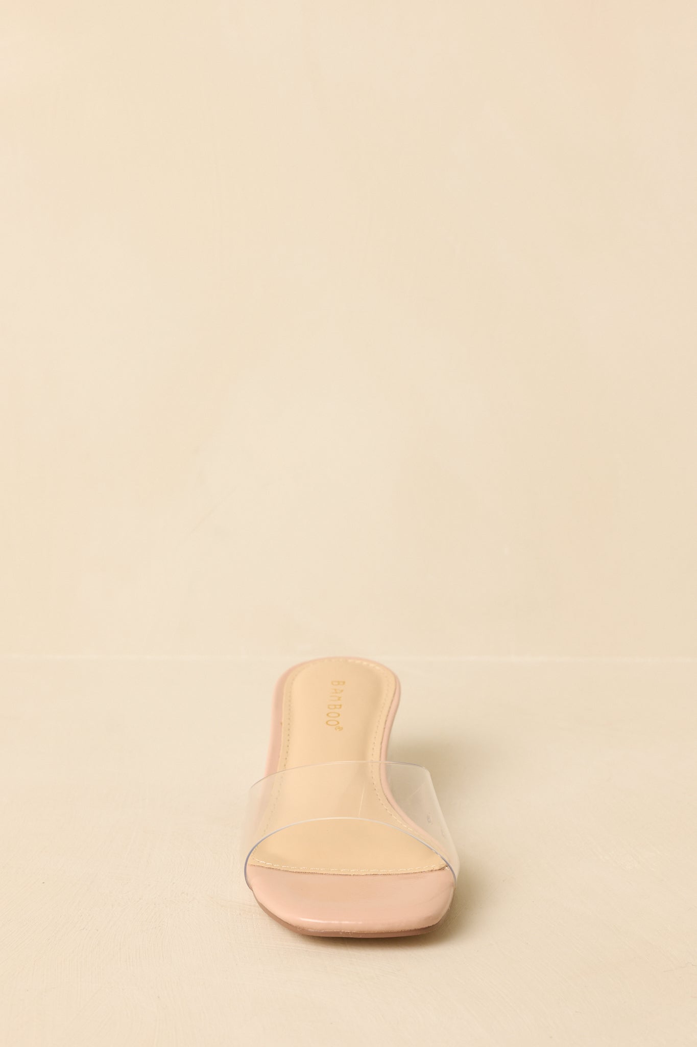 Front-facing view of beige open-toe slide heels against a beige background, highlighting the clear strap and minimalist design.