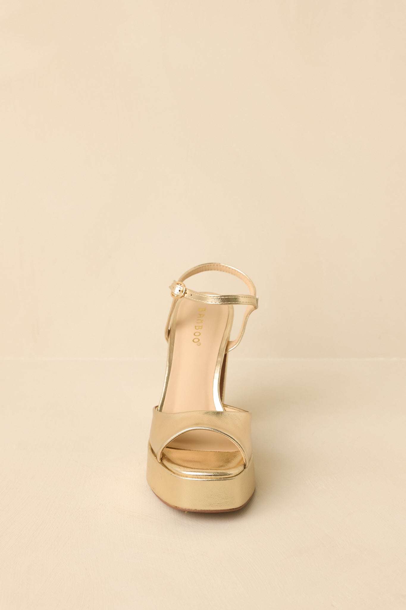 Front view of gold metallic heels on a beige background highlighting the open toe and smooth faux leather finish.