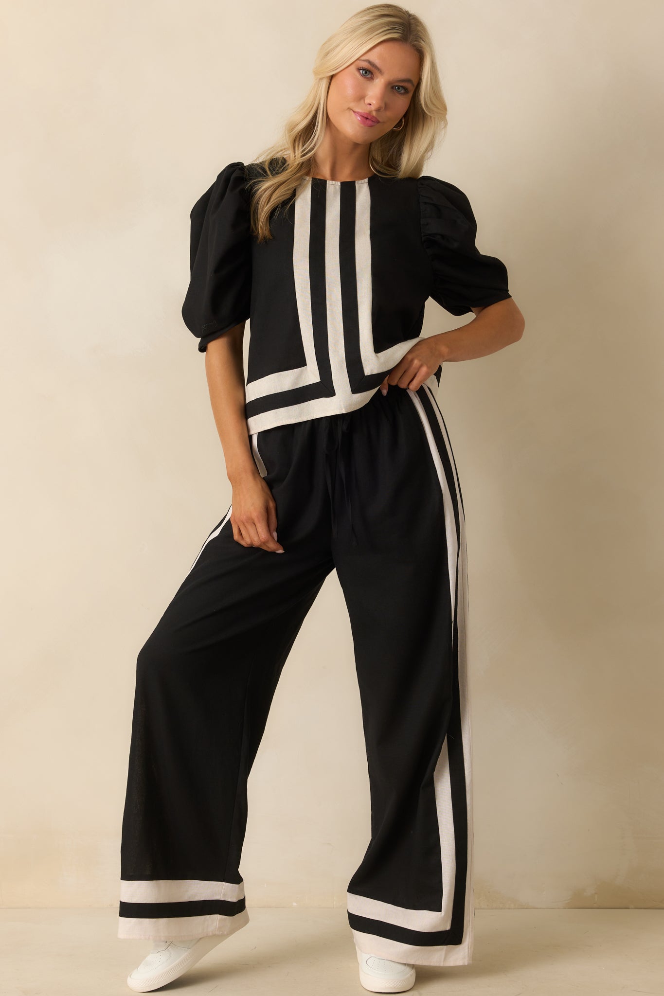 Front-facing view of black wide-leg pants with high-rise elastic waistband, adjustable self-tie drawstring, functional pockets, and ivory and black contrast trim along the legs.
