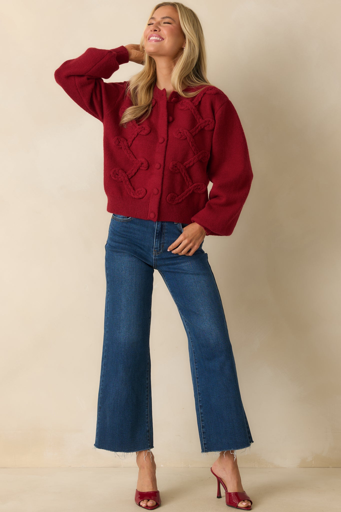 Full-length front view of red knit cardigan featuring long sleeves, front buttons, ribbed trim, and bold braided appliqué across the front.