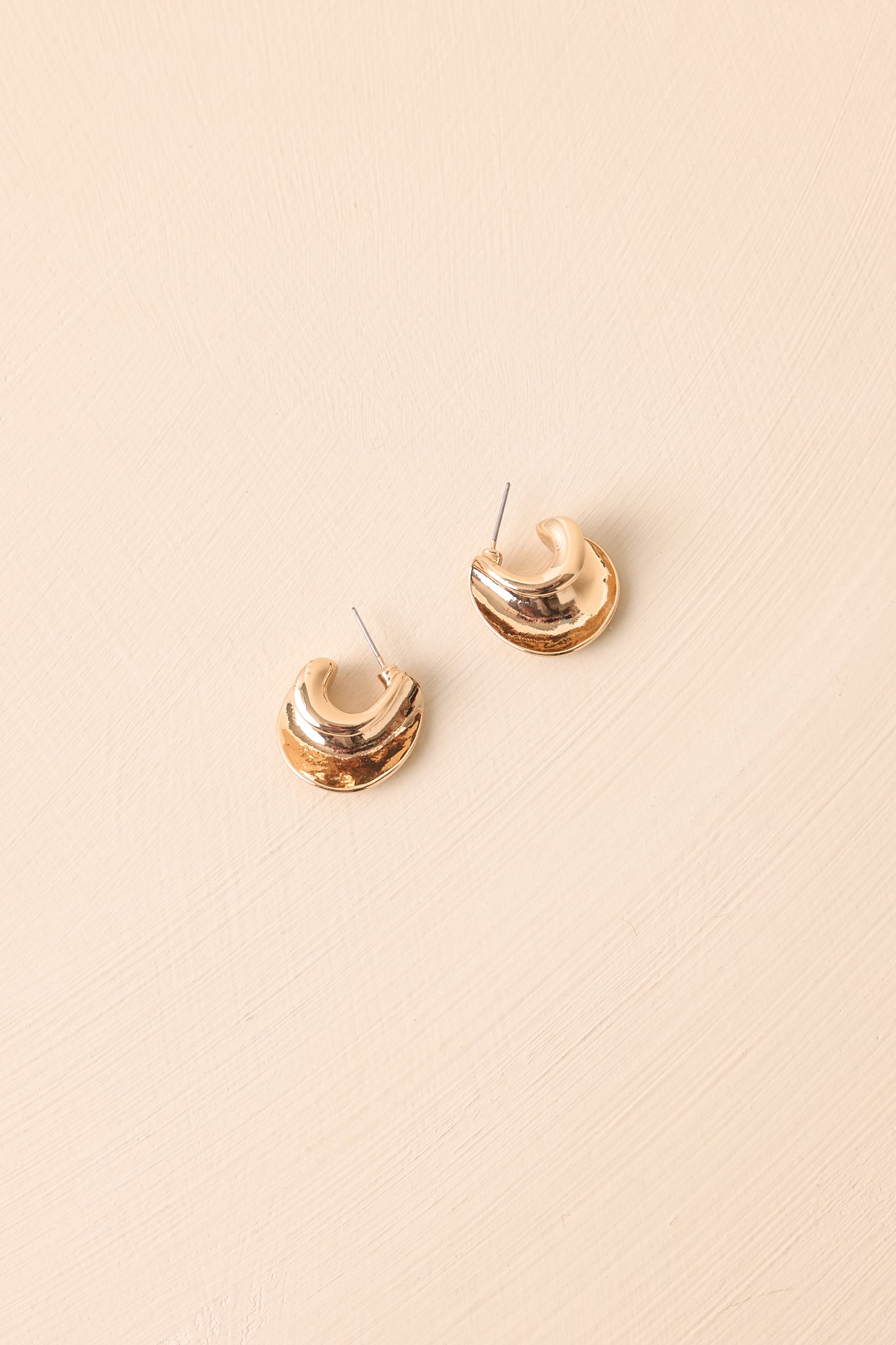 The gold sculptural c-hoop earrings shown against a beige backdrop featuring an open curved silhouette.