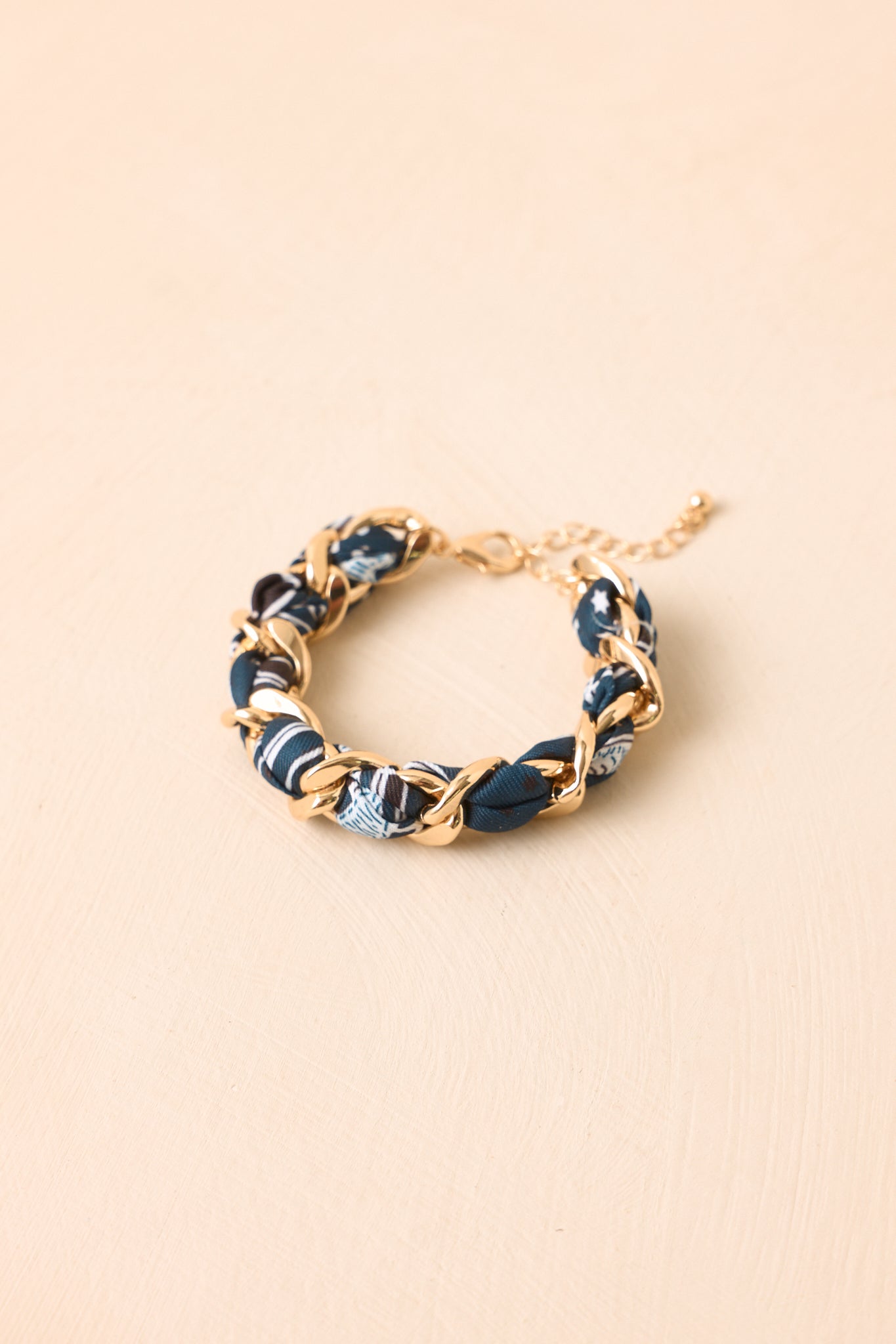 A gold chain bracelet featuring woven segments of navy blue, light blue, and white patterned fabric, displayed on a light beige textured background.