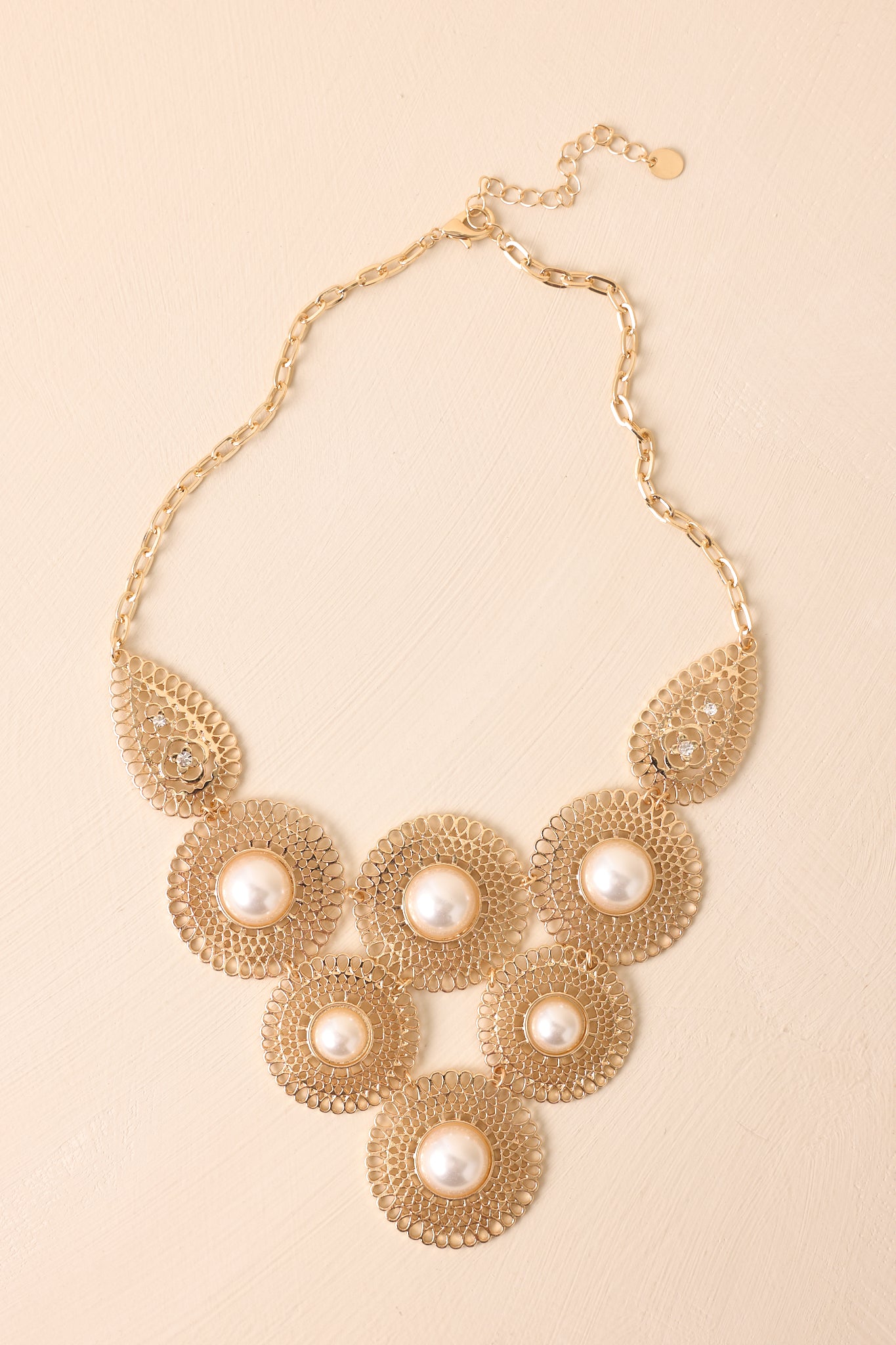 Statement necklace displayed on a beige background, showing the gold chain, filigree discs, and lobster clasp closure.