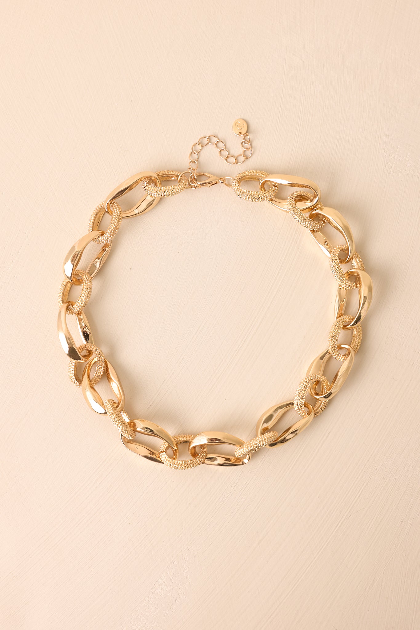 Oversized gold-tone chain necklace displayed on a beige background, showing the polished and textured links along the chain.