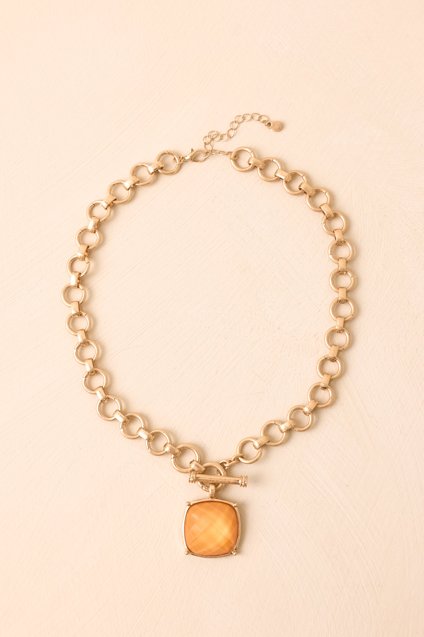 Gold chain necklace displayed against a beige background, highlighting the orange square stone and toggle clasp detail.