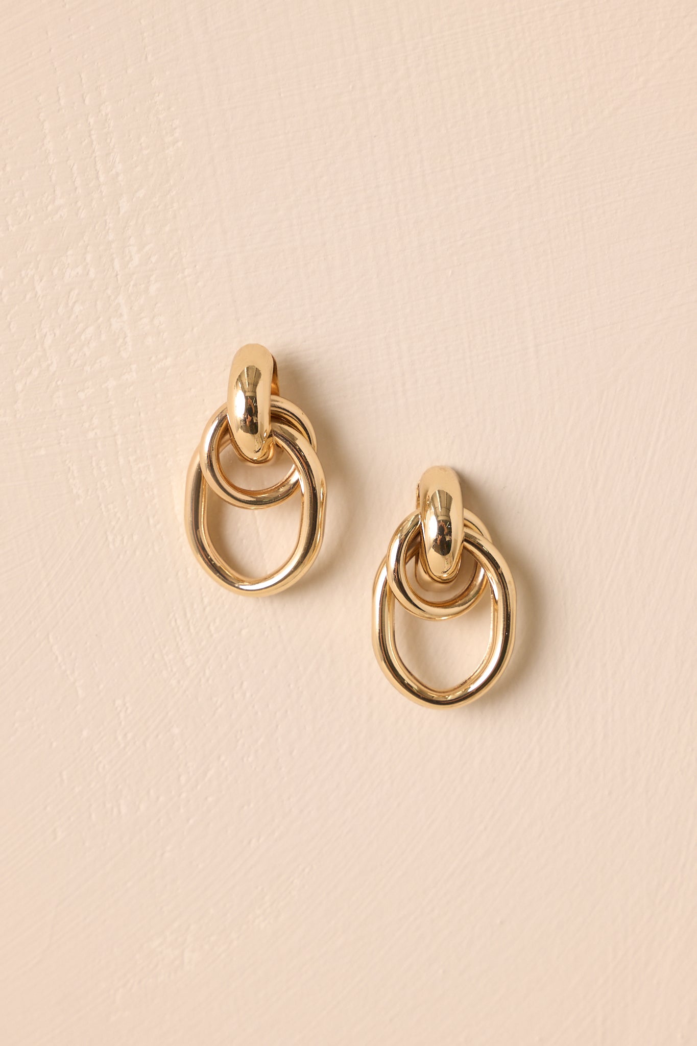 Gold-colored double hoop earrings displayed against a beige background with soft lighting.